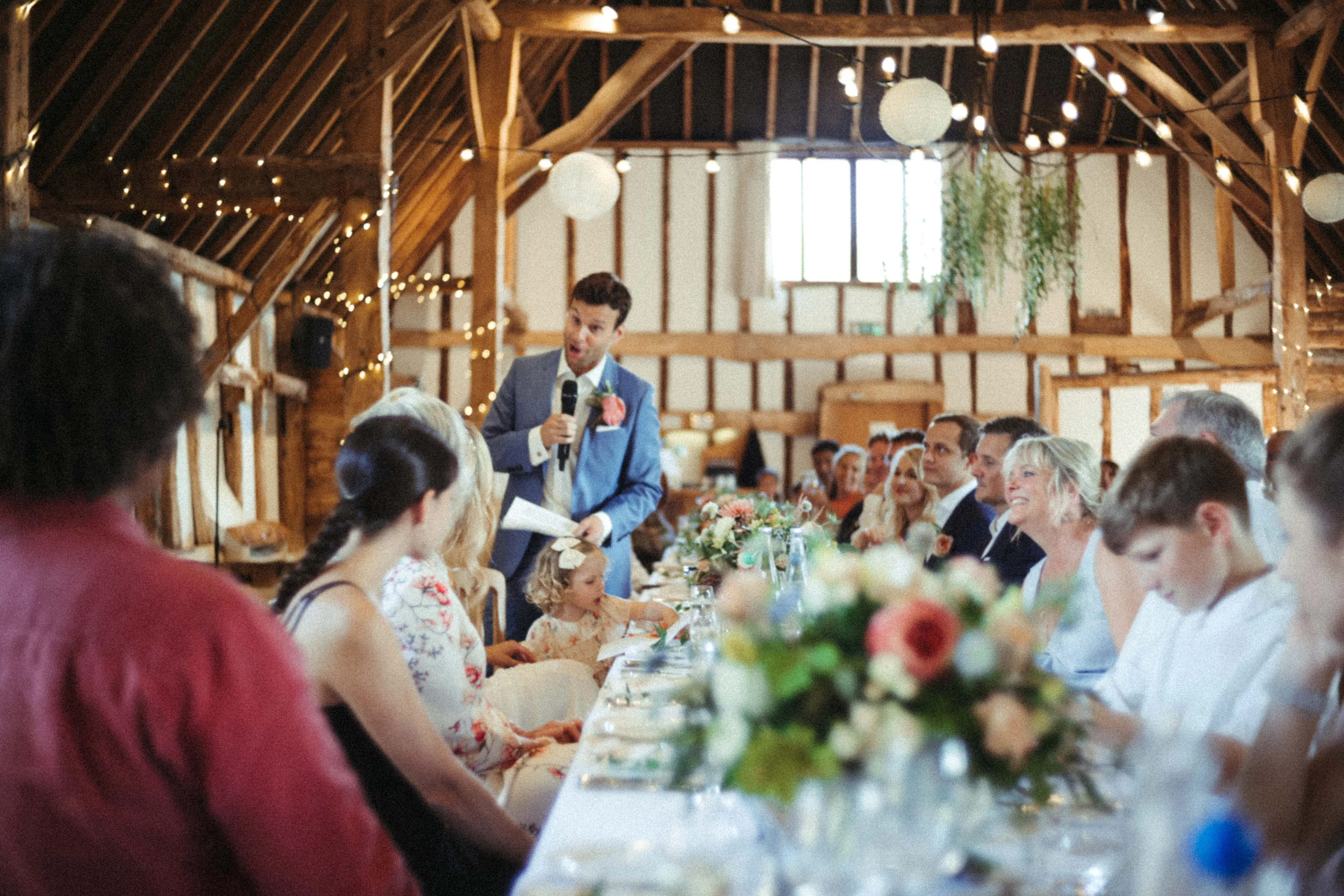 Man in blue suit giving a speech at a barn wedding reception.
