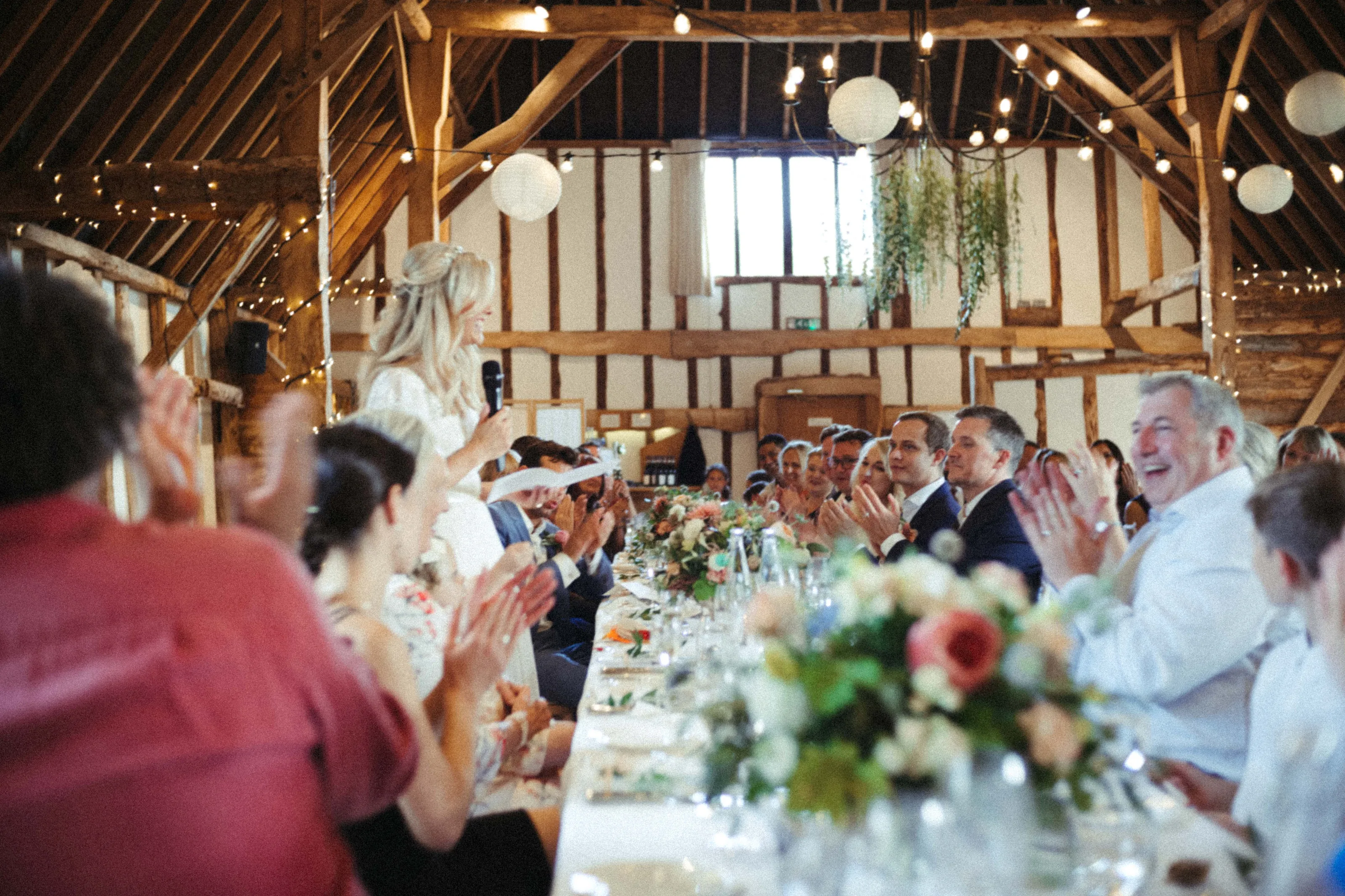 Woman gives speech at barn wedding, guests clap at decorated table.