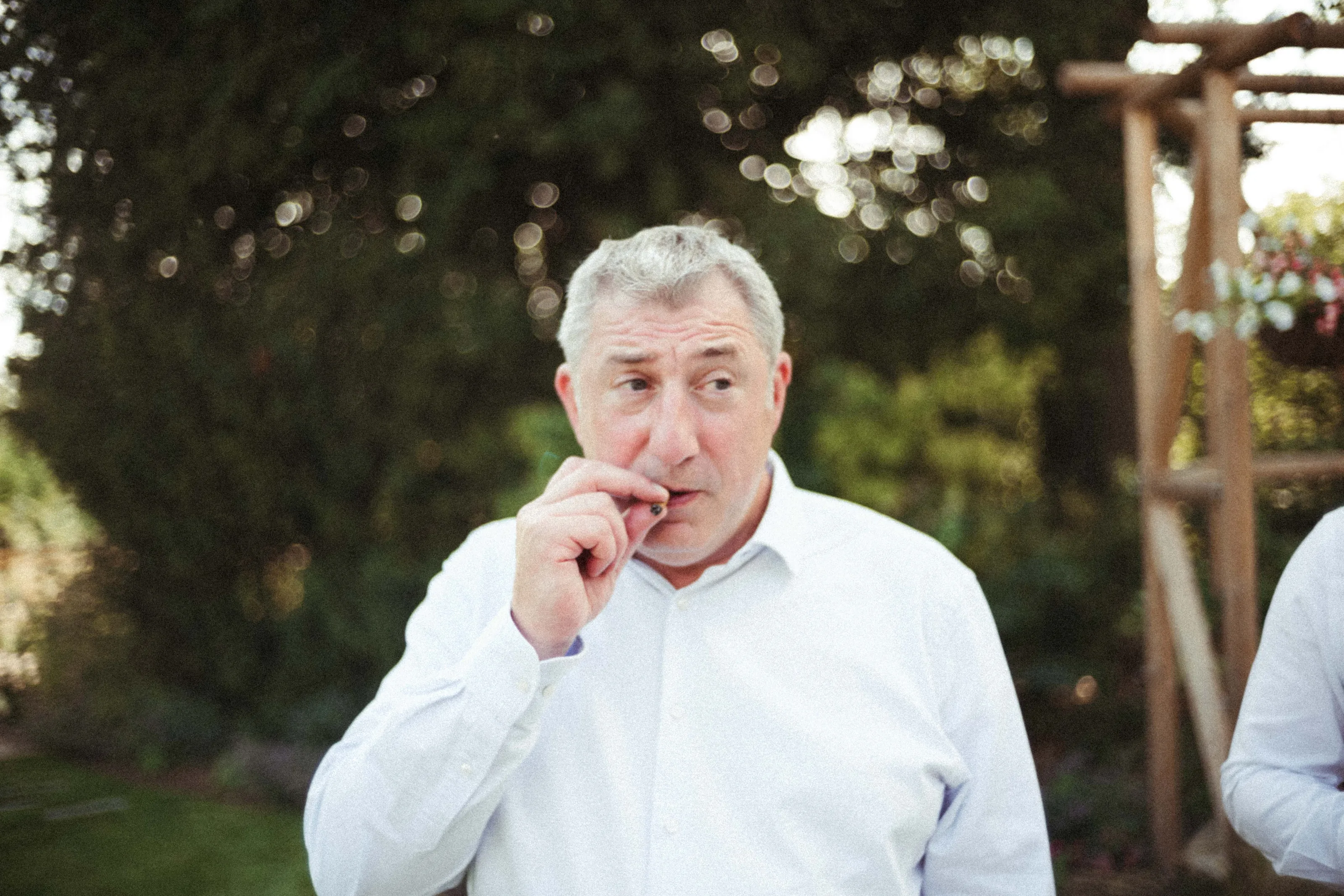 Grey-haired man in white shirt smoking, looking sideways in a garden.