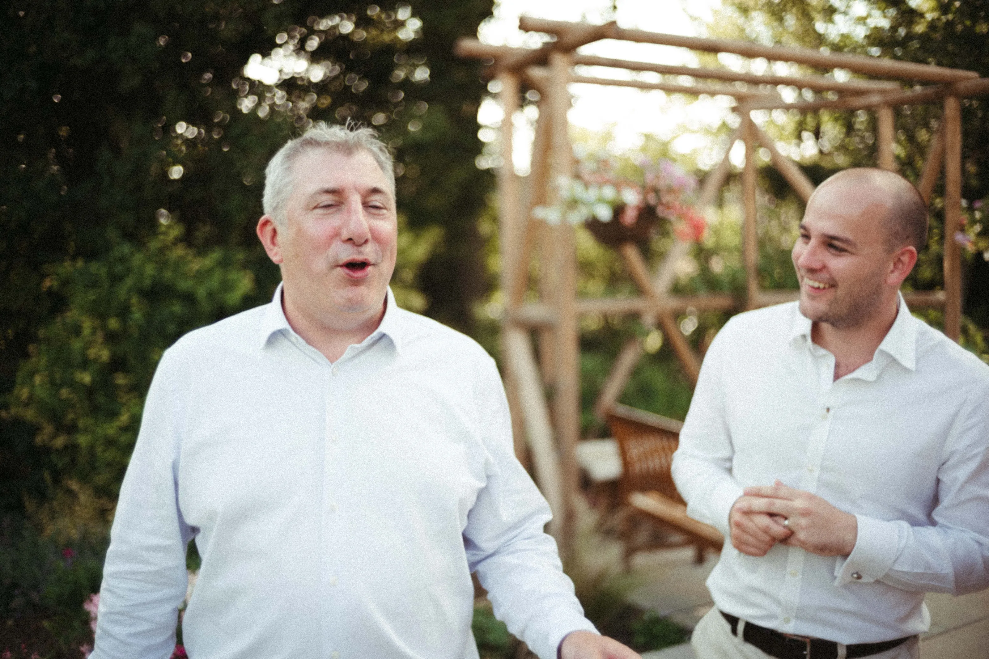 Two men in white shirts talking and smiling in a garden.