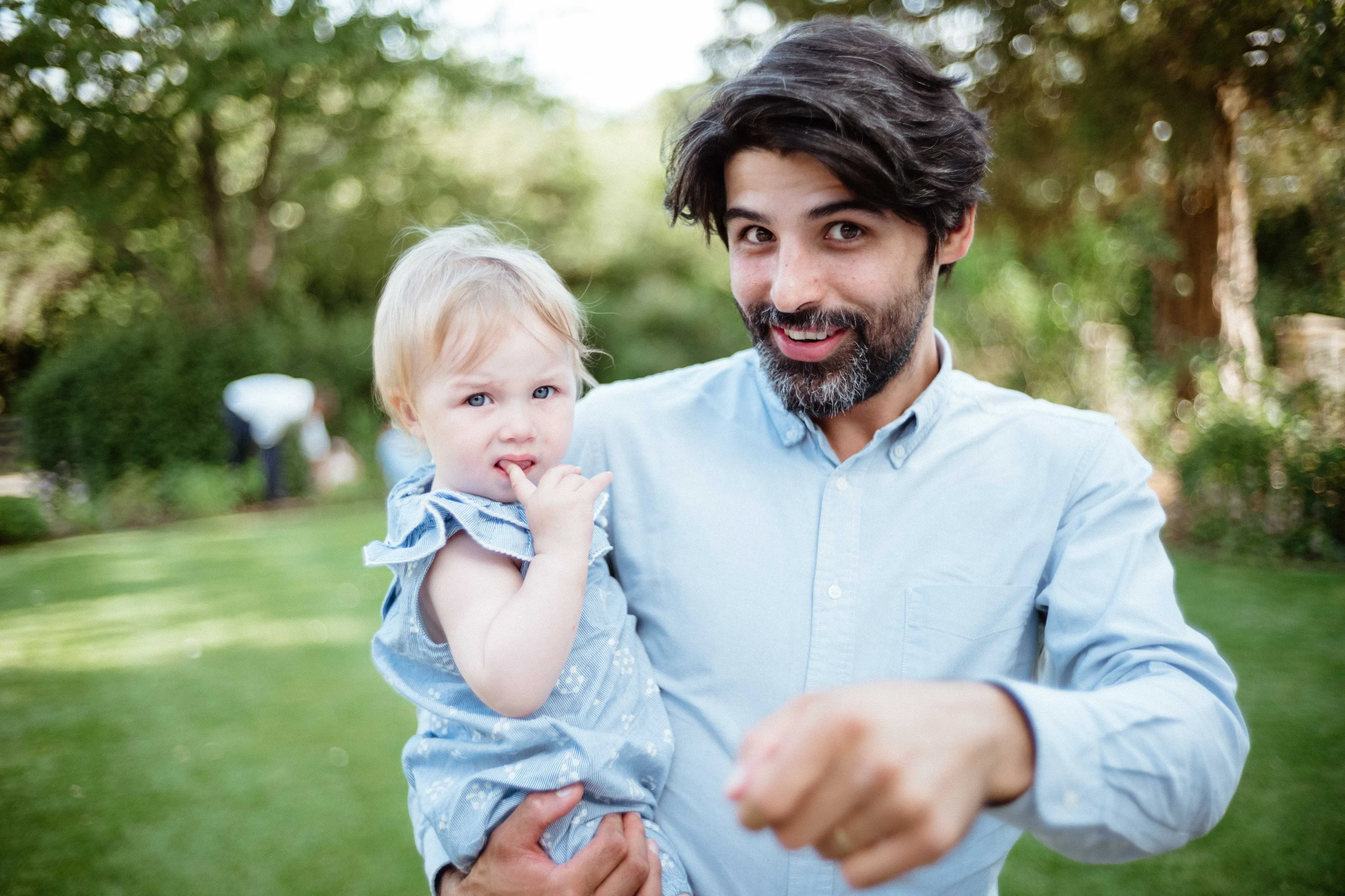Man holding blonde toddler, pointing at camera, outdoors.