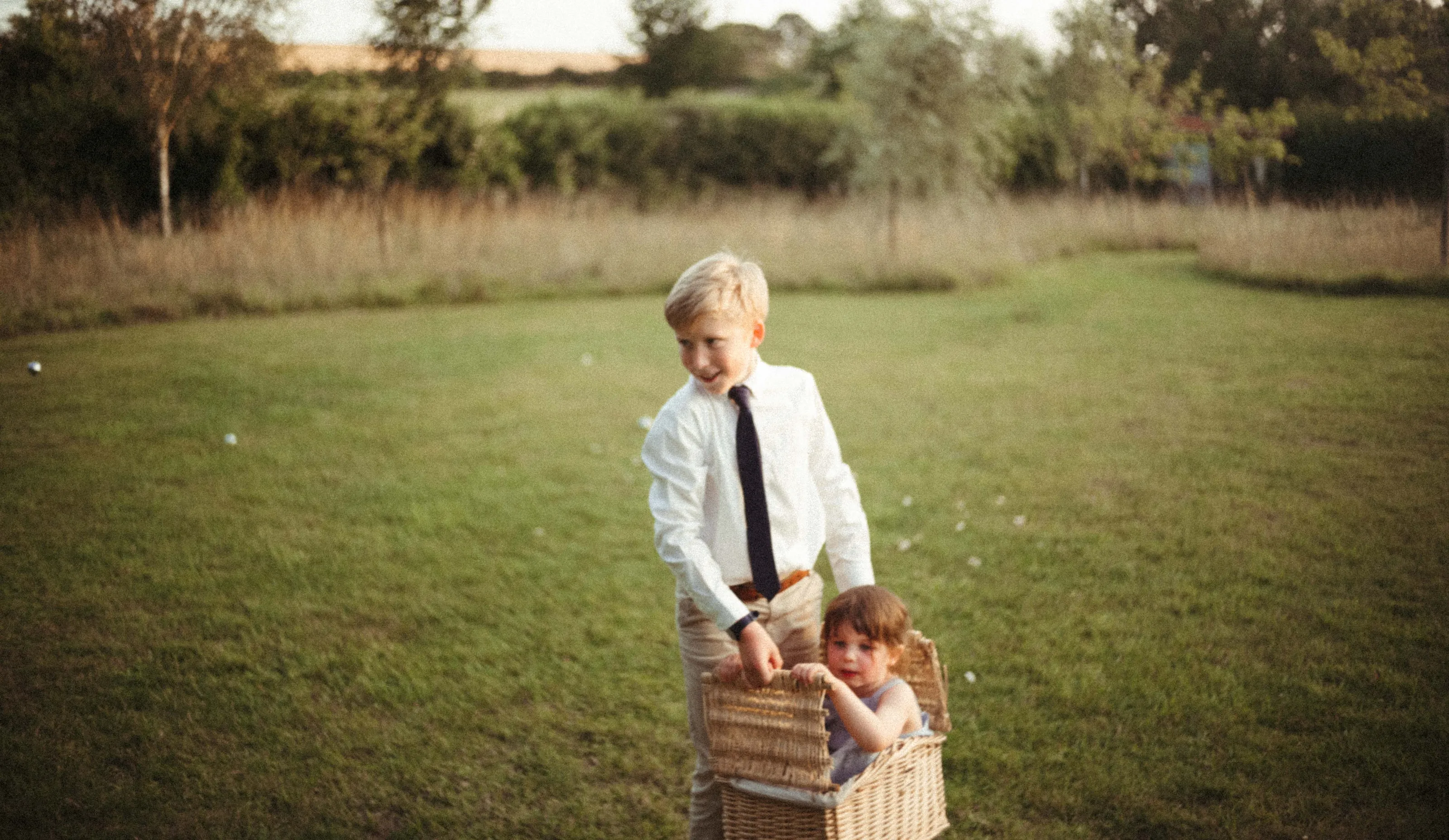 Smiling boy pulls toddler in wicker basket across a green field.