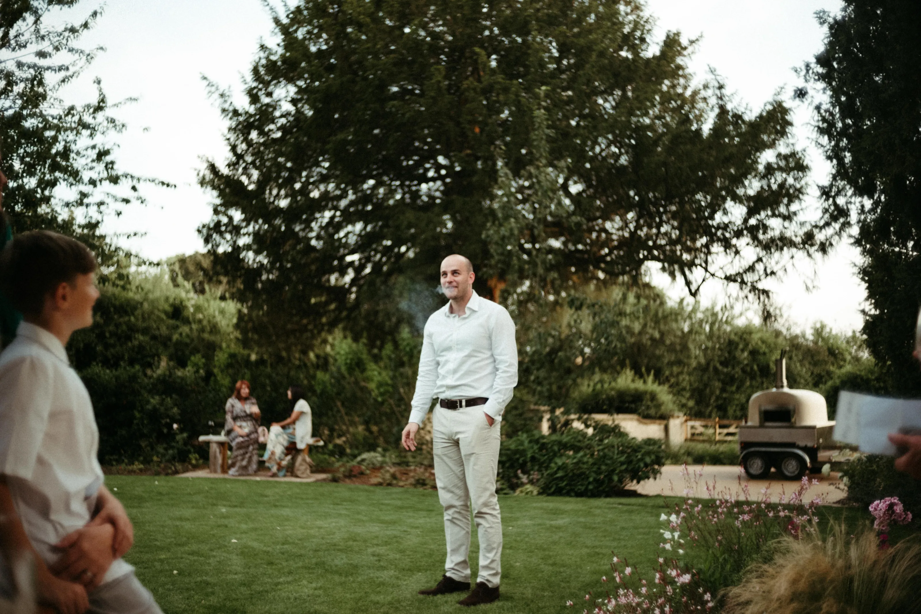 Smiling man with smoke, boy, and mobile pizza oven on lawn.