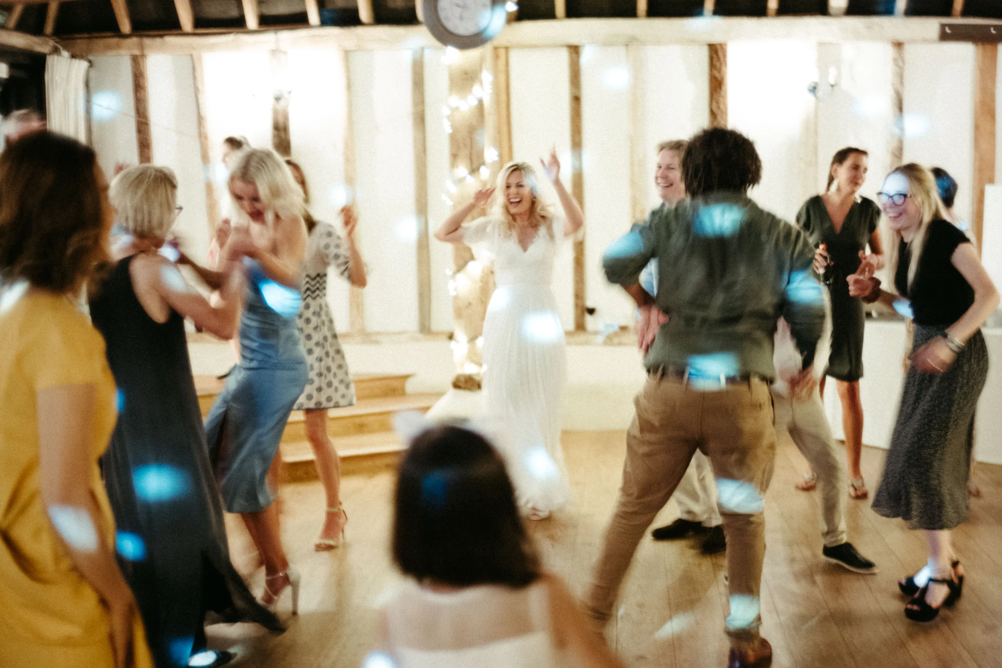 People dancing and laughing, a woman in white dress, with string lights and blue reflections.