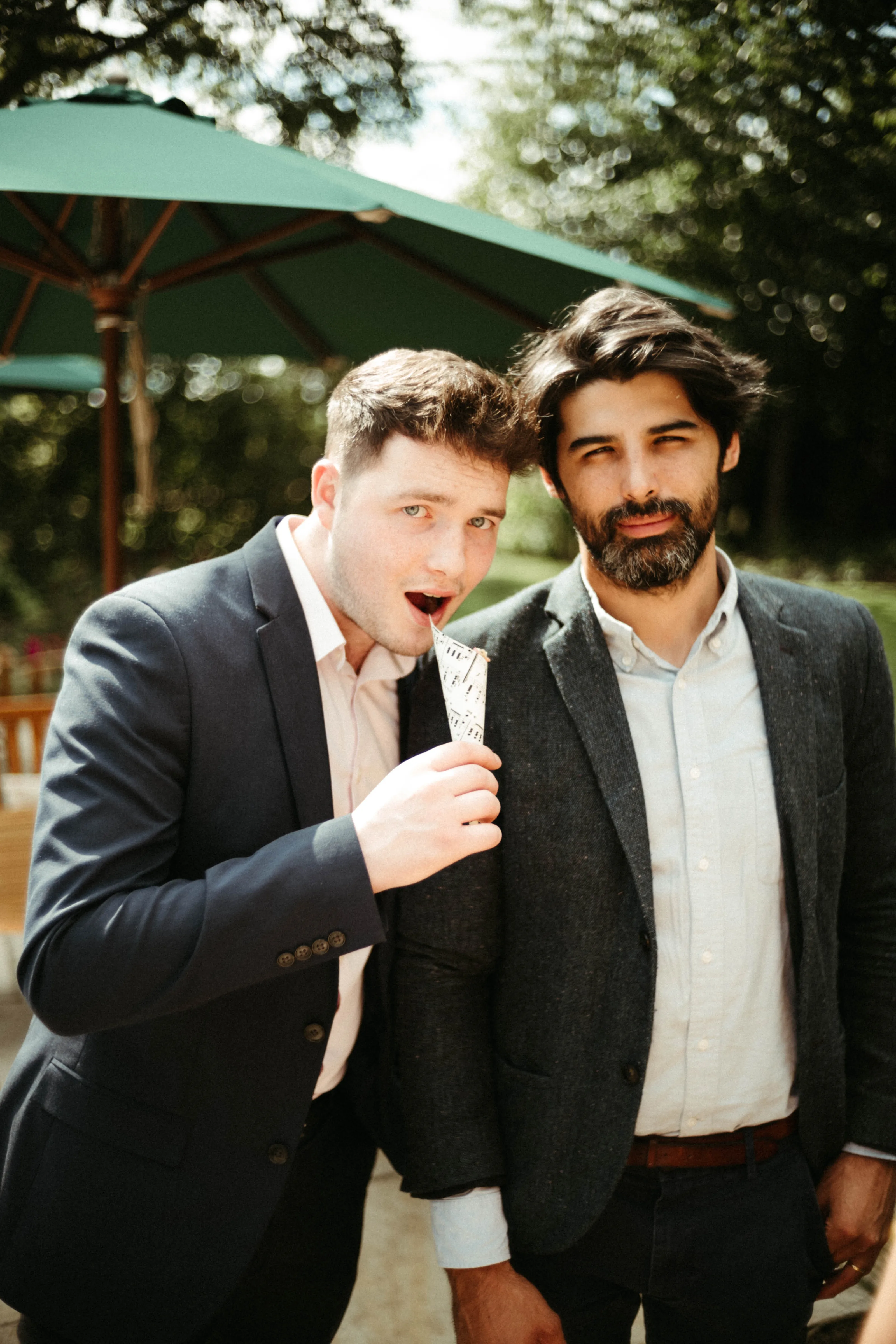 Two men outdoors, one with paper in mouth, the other bearded.