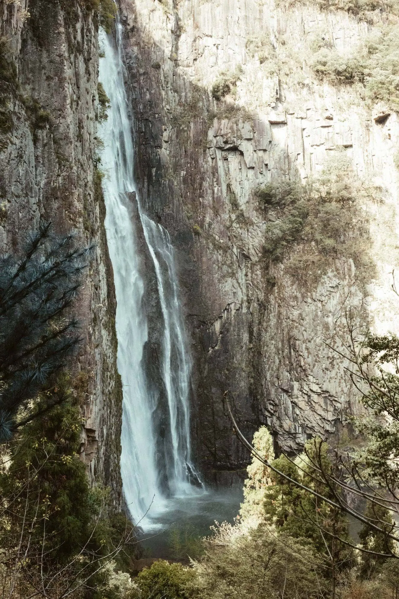 A waterfall dropping in a single thread down a sheer, weathered cliff, greenery clinging to the rock.