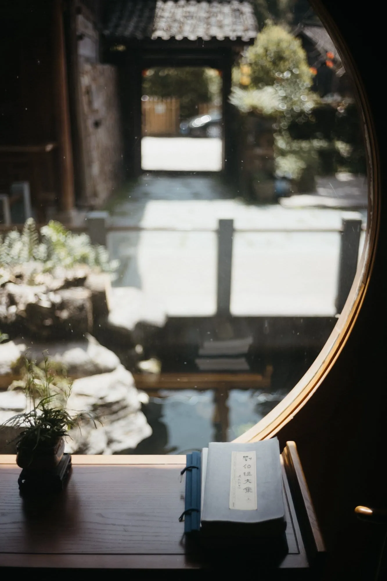 A blue thread-bound Chinese book on a wooden table beside a circular window, a rockery garden reflected in the glass.