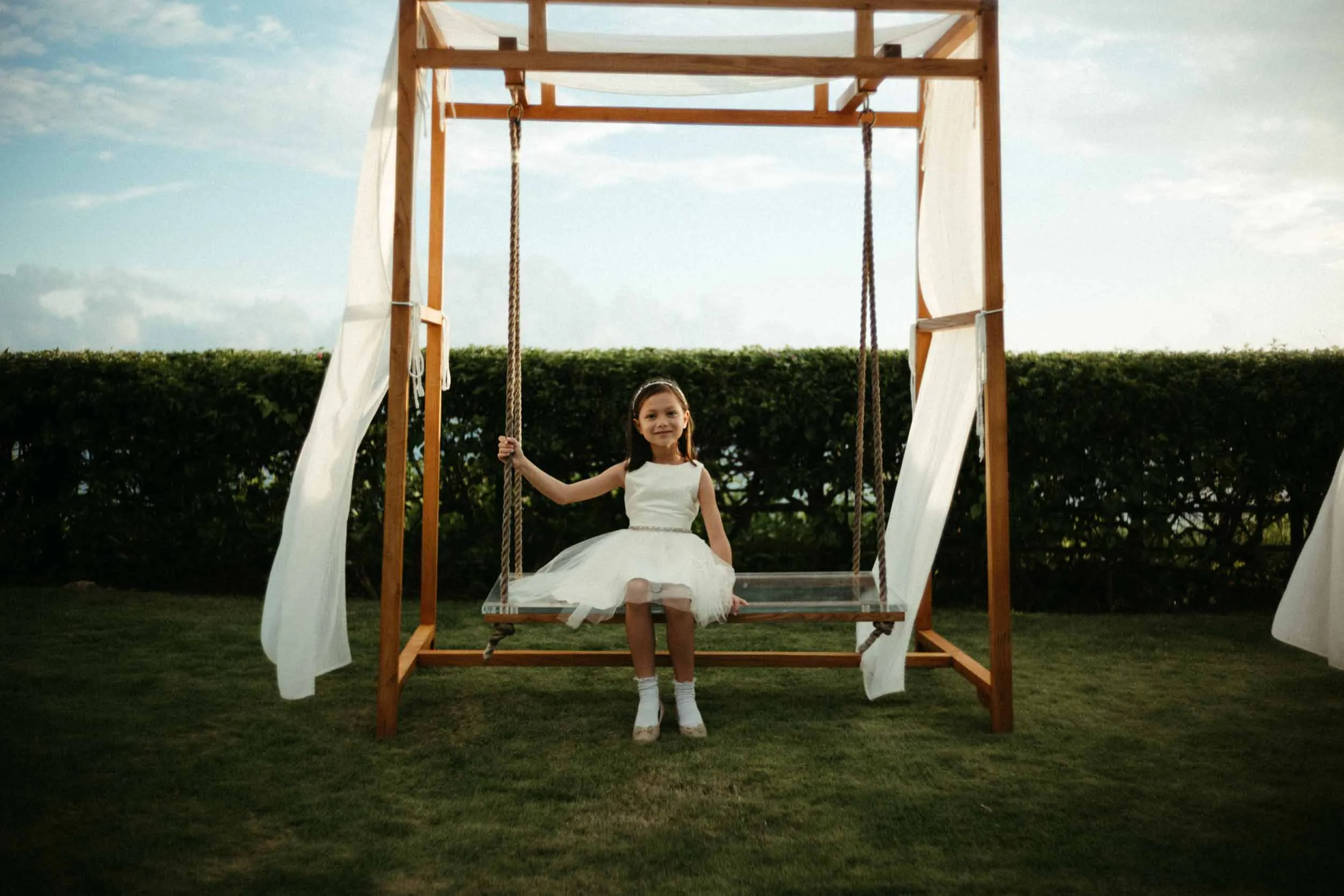 Smiling girl in white dress on a swing within a draped wooden frame.