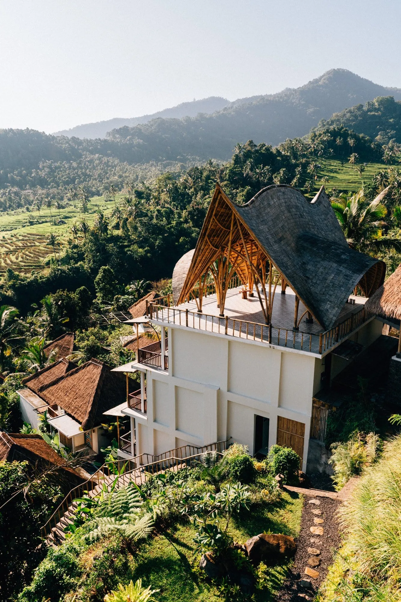 A bamboo-framed villa with a curved shingled roof rising above thatched huts, terraced rice fields and forested mountains behind.