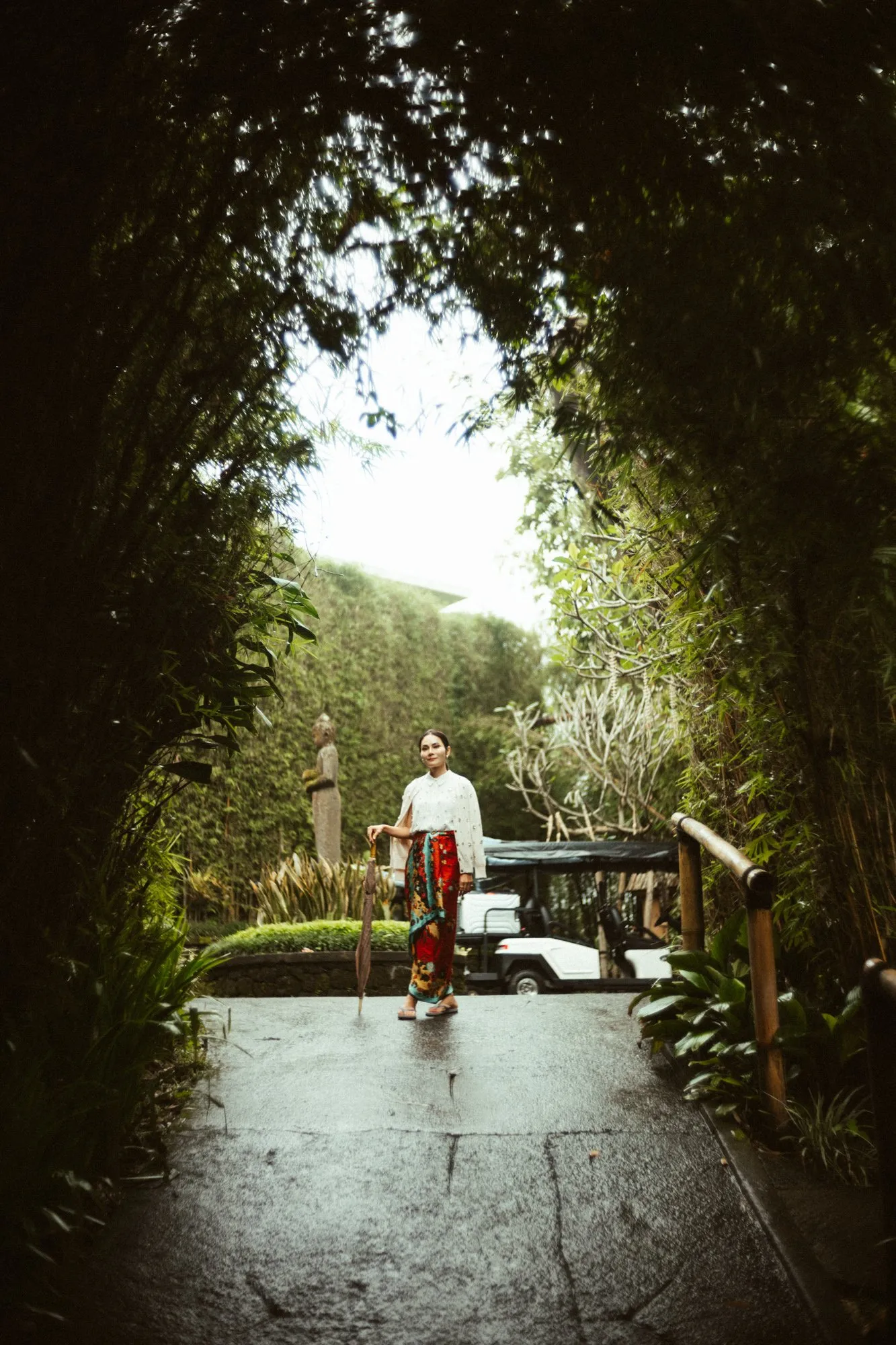 A woman in traditional Balinese dress holding an umbrella on a path through a lush garden.