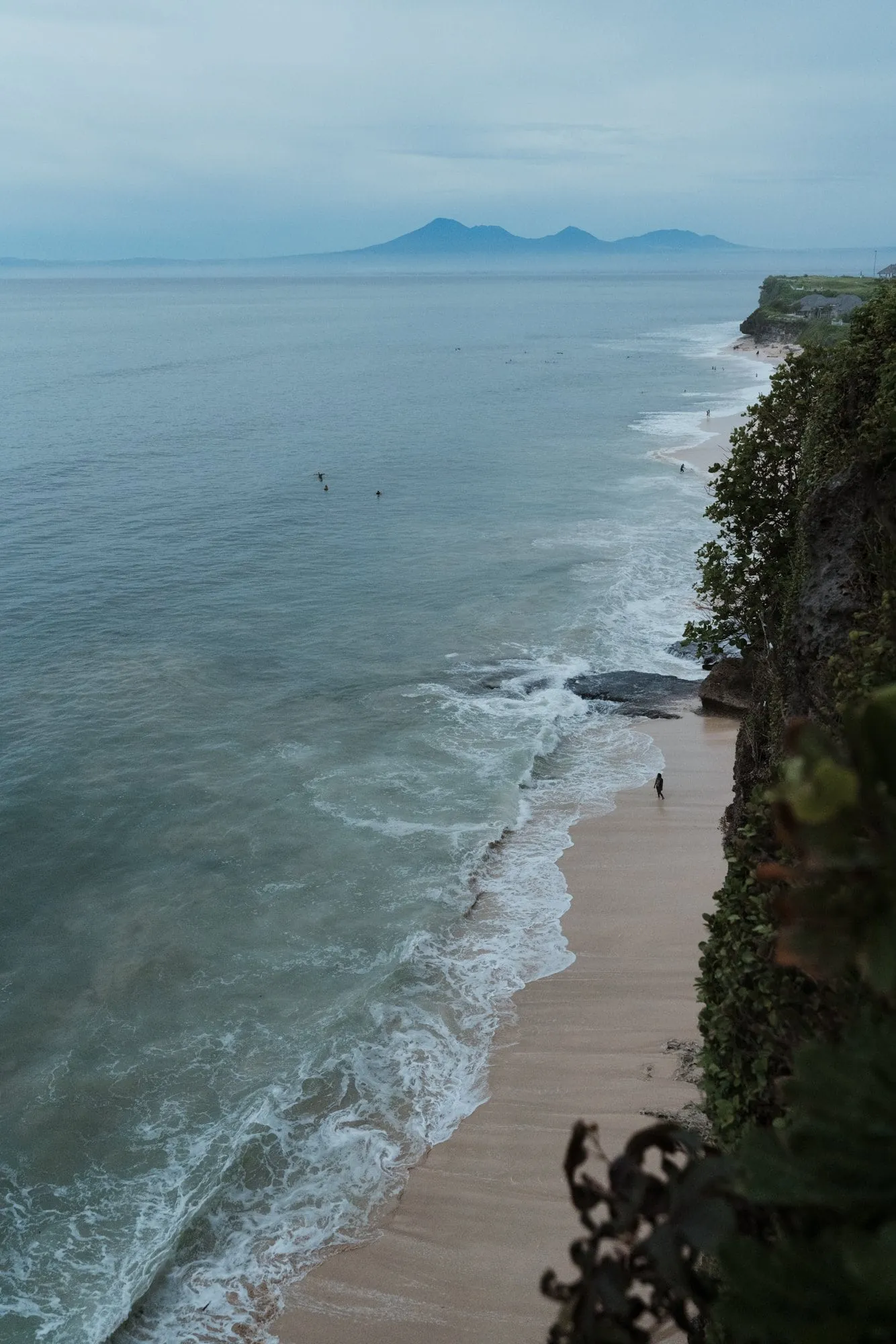 A view from the cliff down to a narrow beach and breaking surf, a lone figure on the sand, a hazy volcano across the bay.
