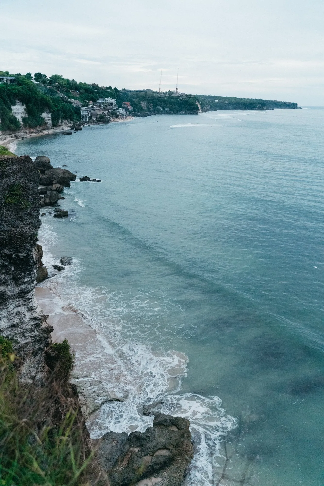 A clifftop view along a curving coastline of white limestone cliffs and turquoise sea, a village and two masts on the far headland.