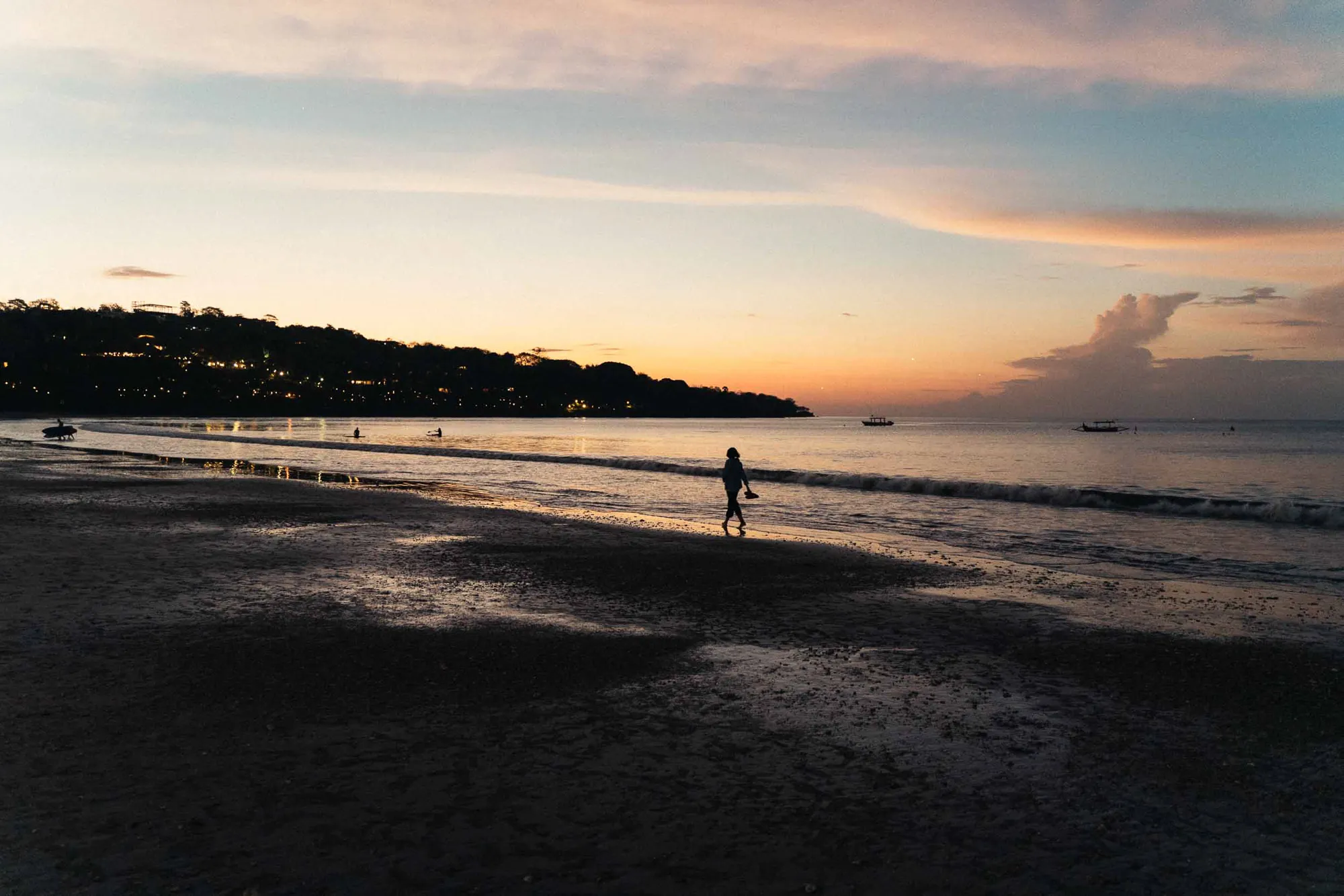 Dusk beach with person walking, waves, boats, and distant illuminated buildings.