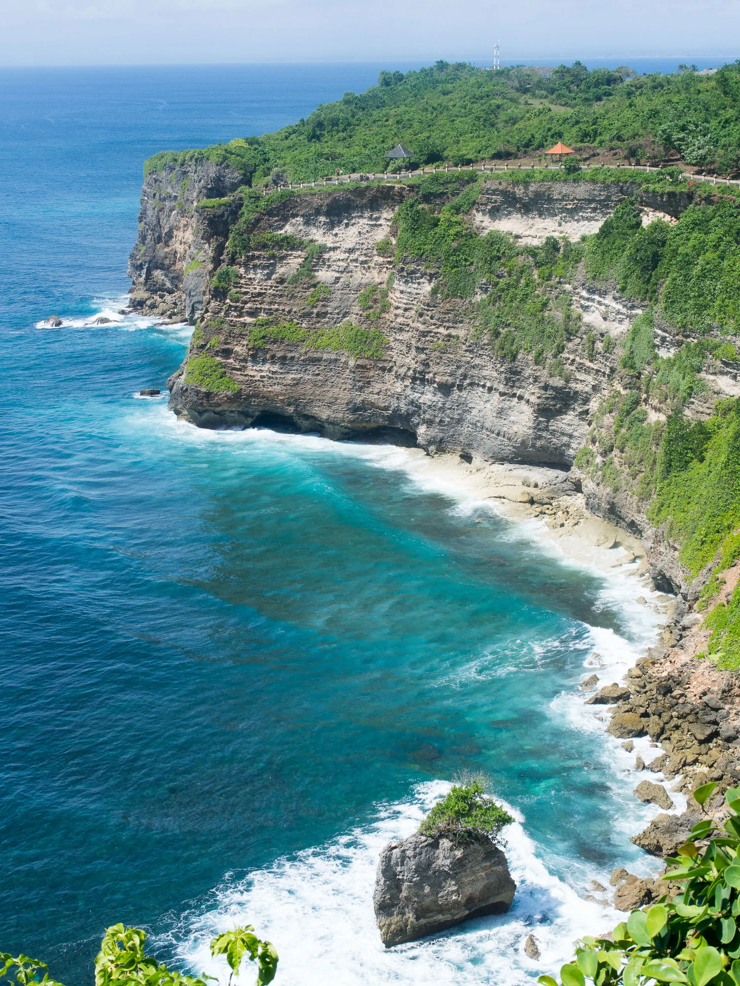 Coastal cliff with green vegetation, turquoise sea, and white waves.