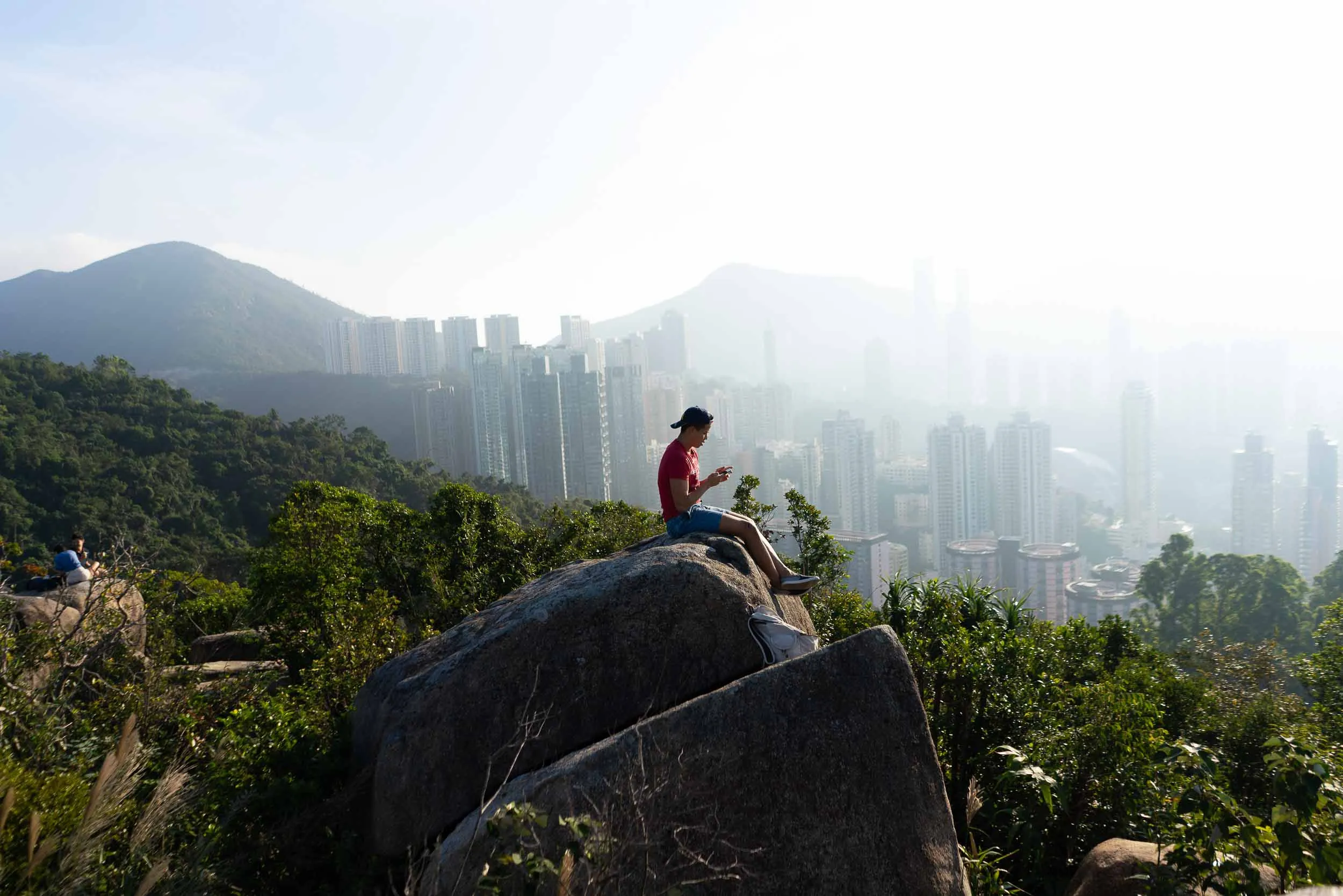 Man on rock using phone, overlooking hazy city and mountains.