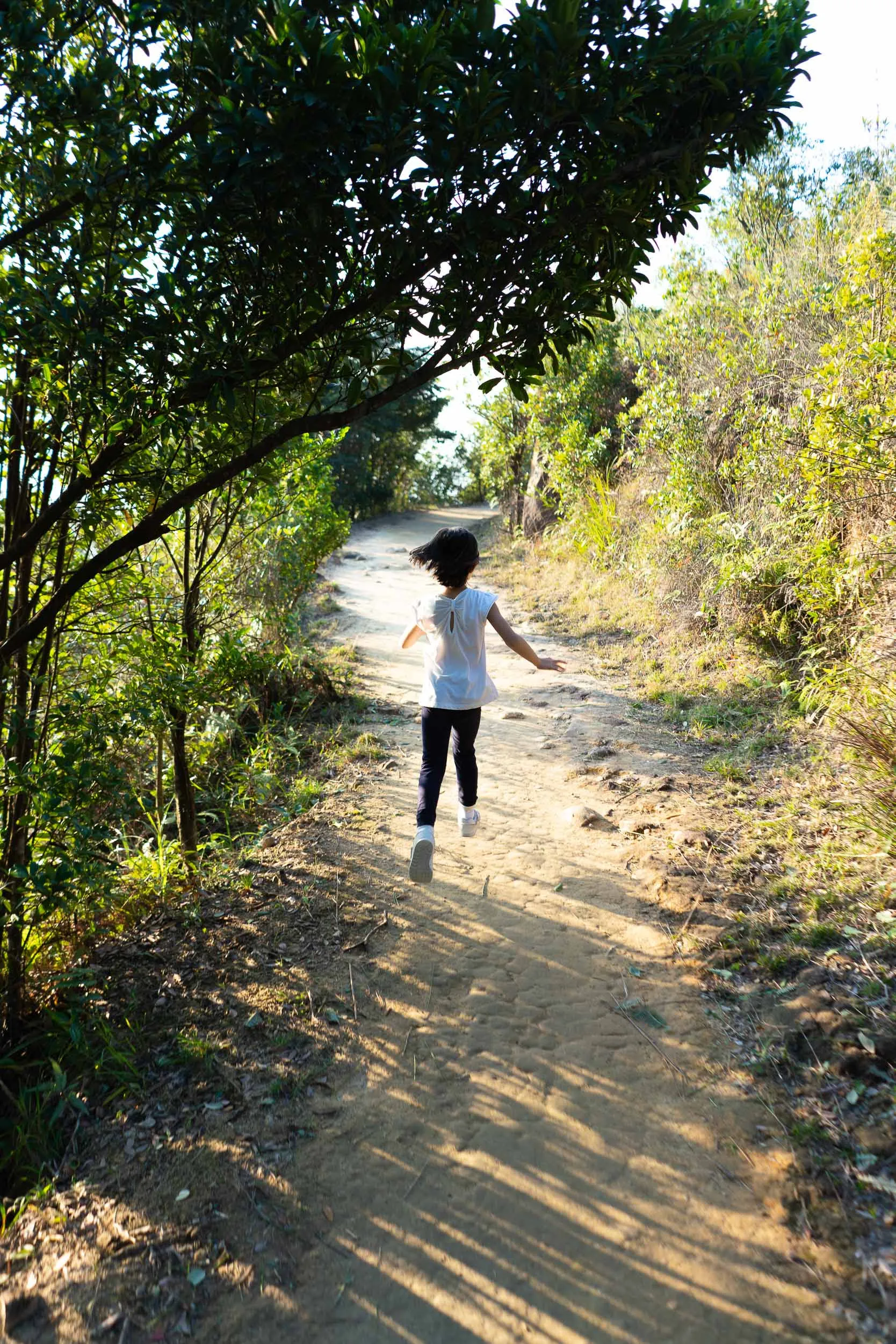 Child running on a sun-dappled dirt path through green trees.