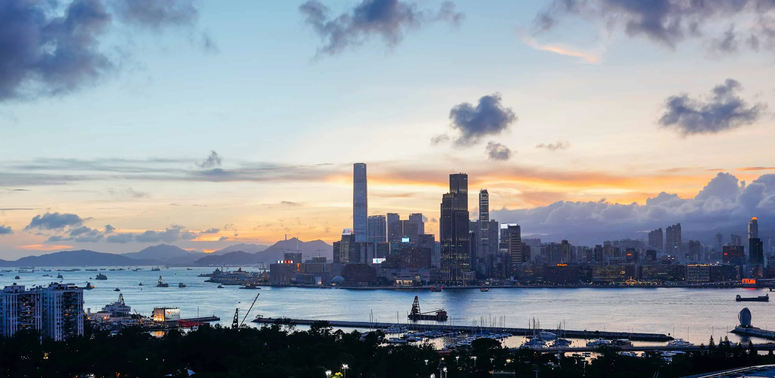 Sunset over a city harbour with skyscrapers, boats, and mountains.