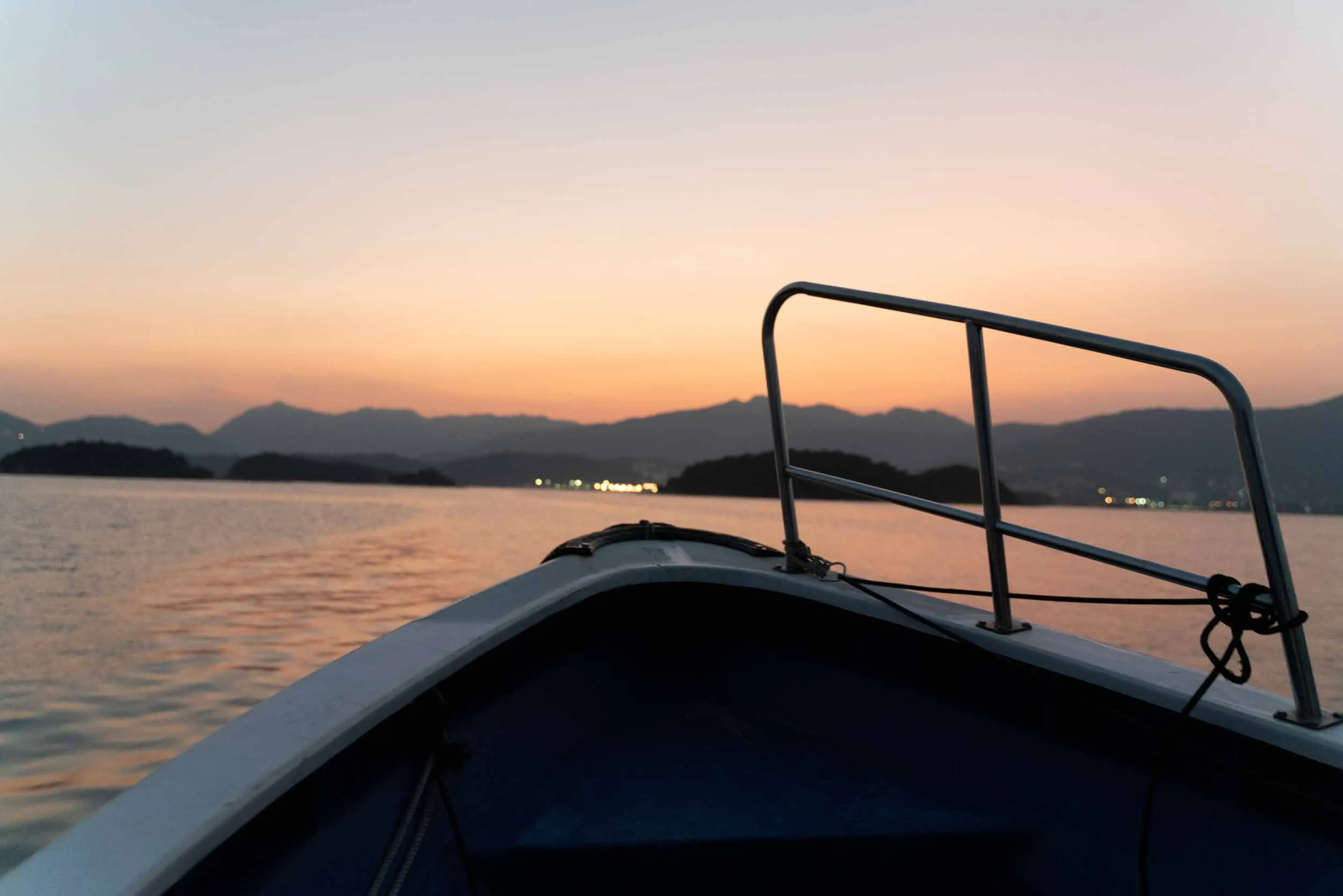 Boat bow on water at sunset with distant mountains and lights.