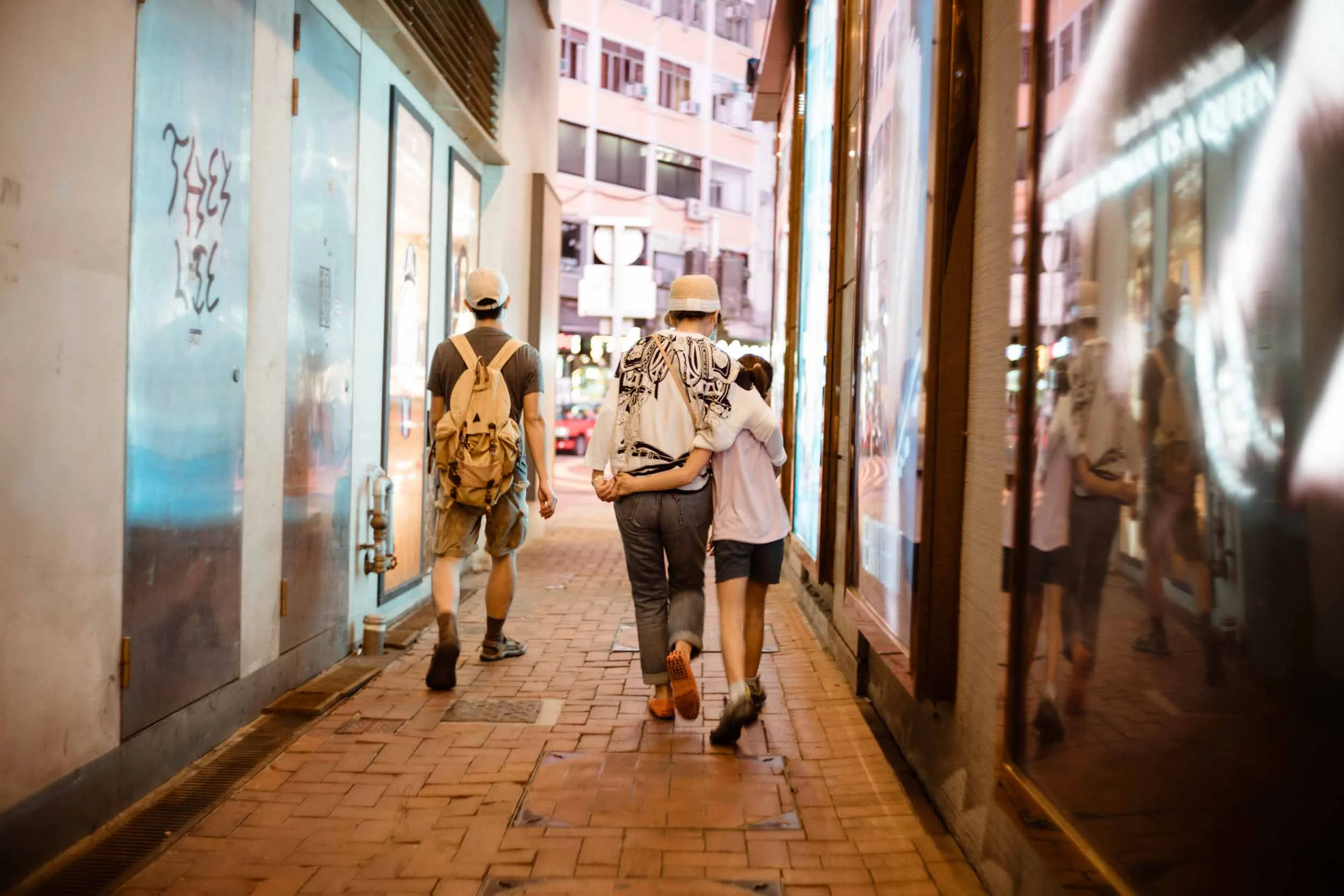 Three people walk along a narrow brick alley past illuminated shop windows.