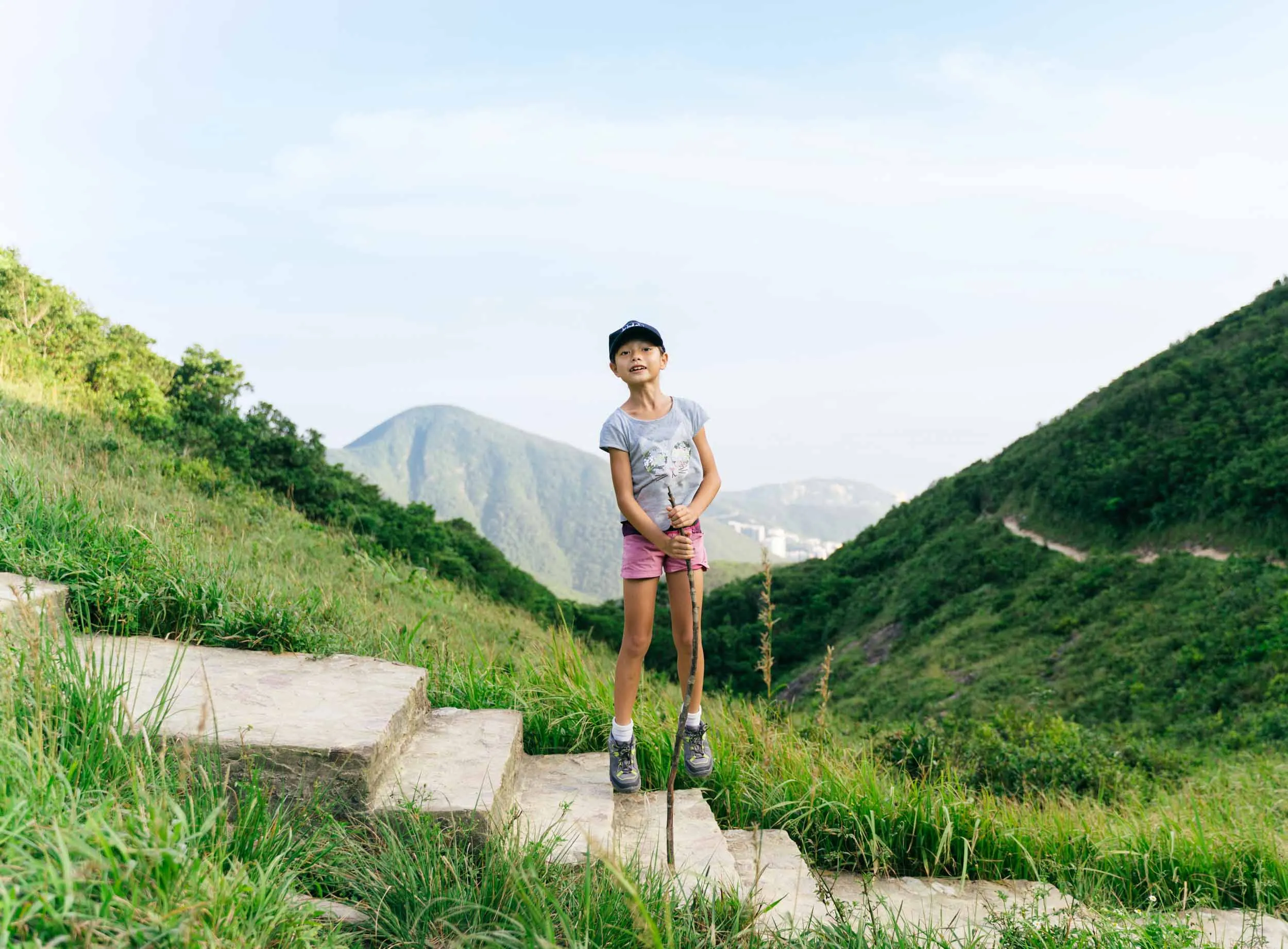 Smiling girl on stone steps, holding a stick, surrounded by green hills.