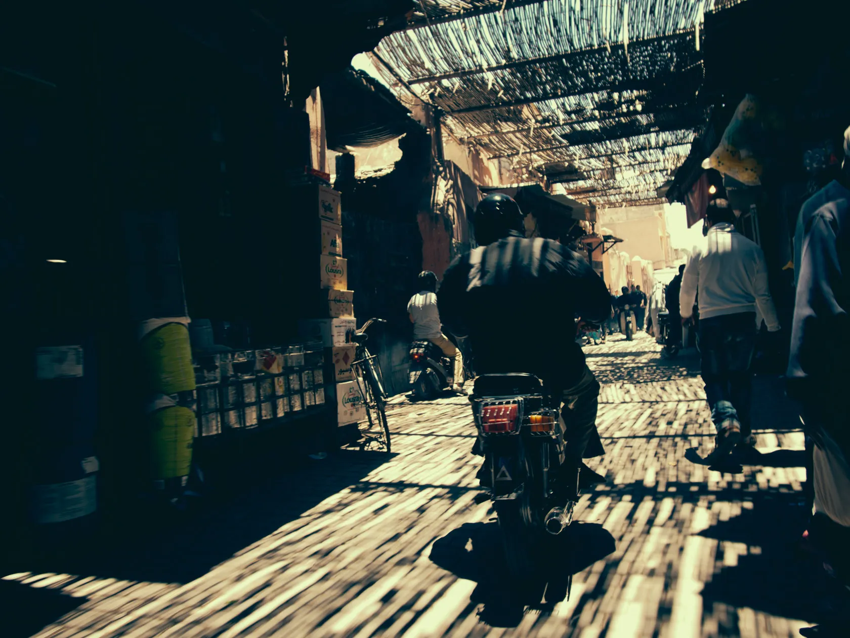 Motorbike rider in a busy, sunlit market street with woven roof.