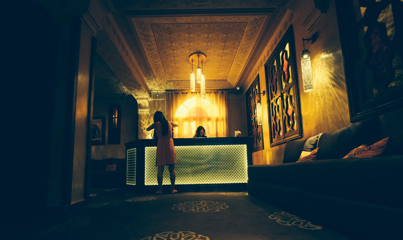 Woman at glowing reception desk in ornate, dimly lit room with patterned ceiling and sofa.