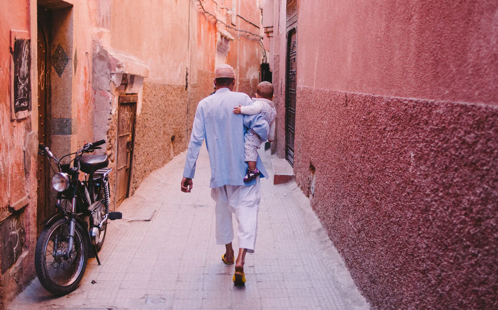 Man carrying child walks down a narrow, reddish alley past a parked moped.