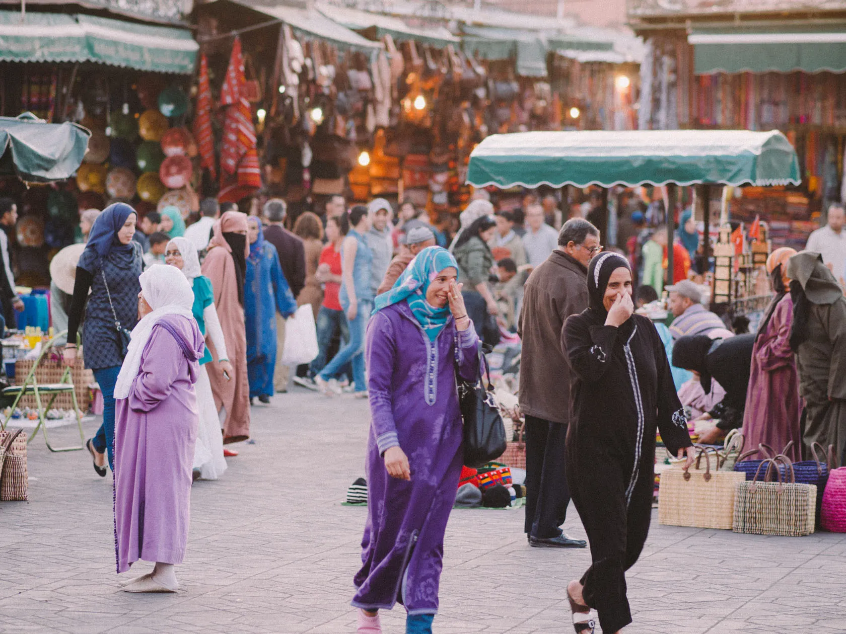 Bustling market scene with people, stalls, and smiling women in hijabs.