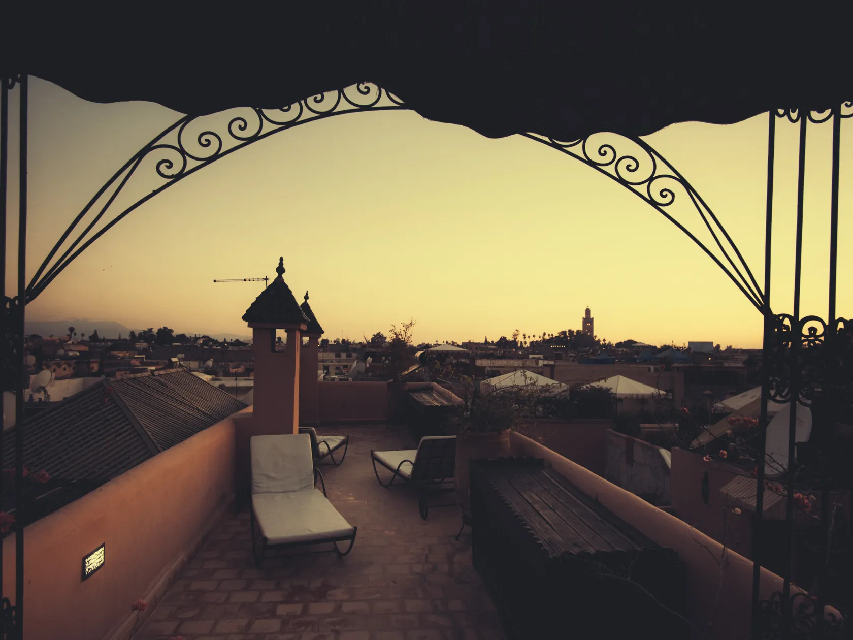 Ornate archway frames rooftop terrace with lounge chairs, city, and minaret at sunset.
