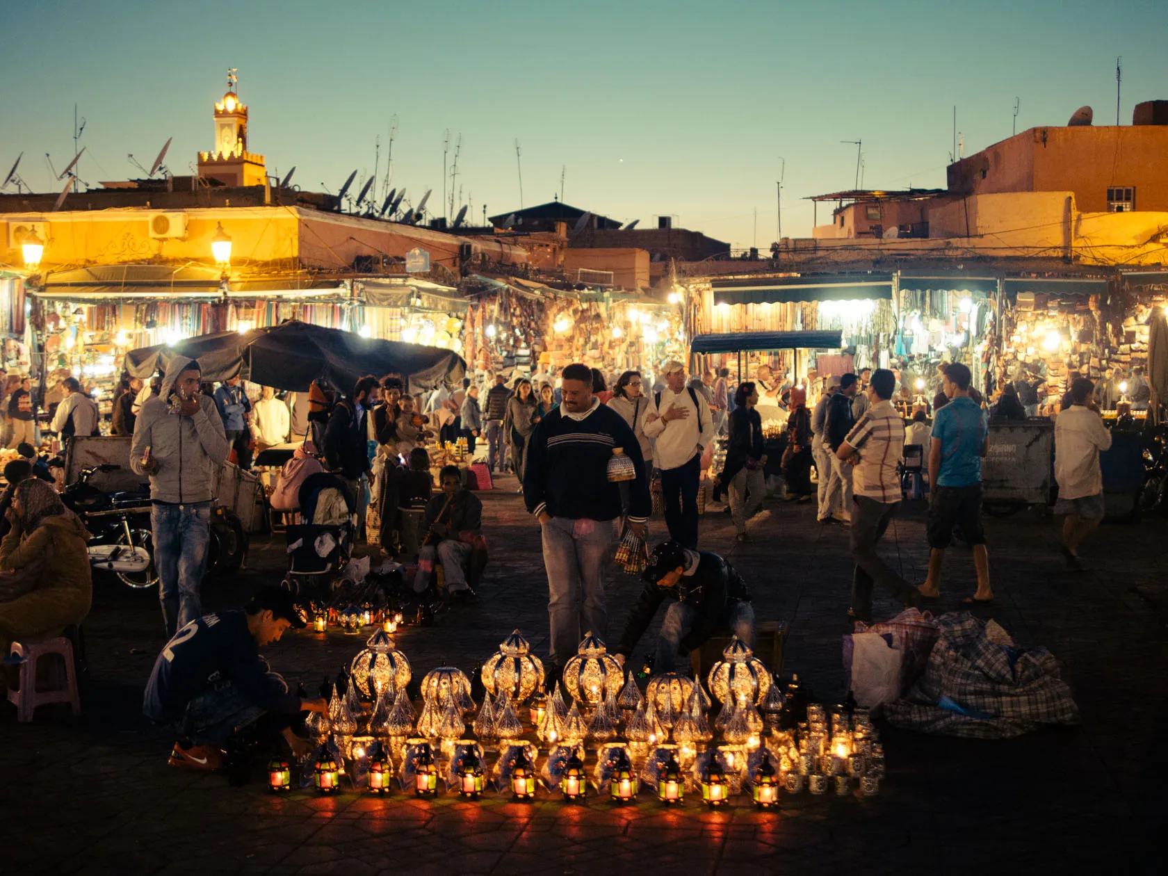 Busy night market with people, illuminated stalls, and glowing lanterns at dusk.