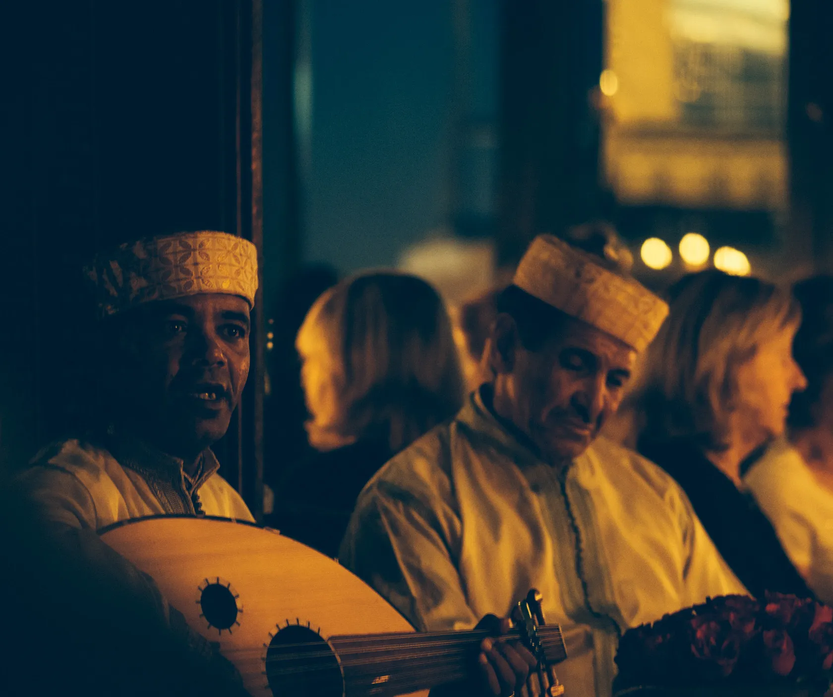 Two men in traditional attire, one playing an oud, in dim light.