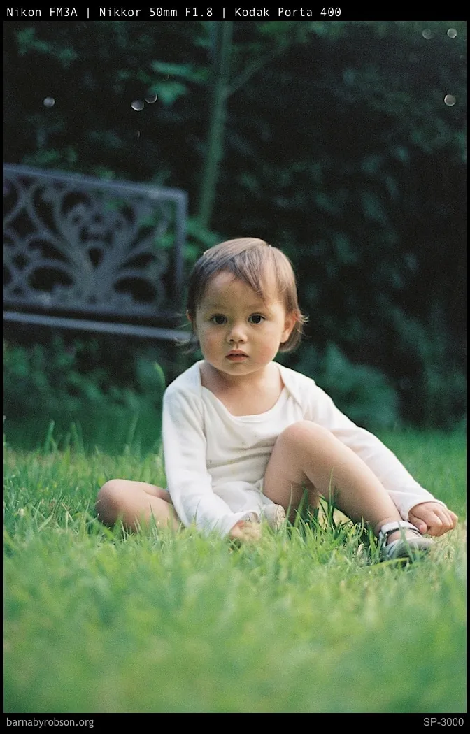 Toddler in cream top sitting on green grass, looking forward.