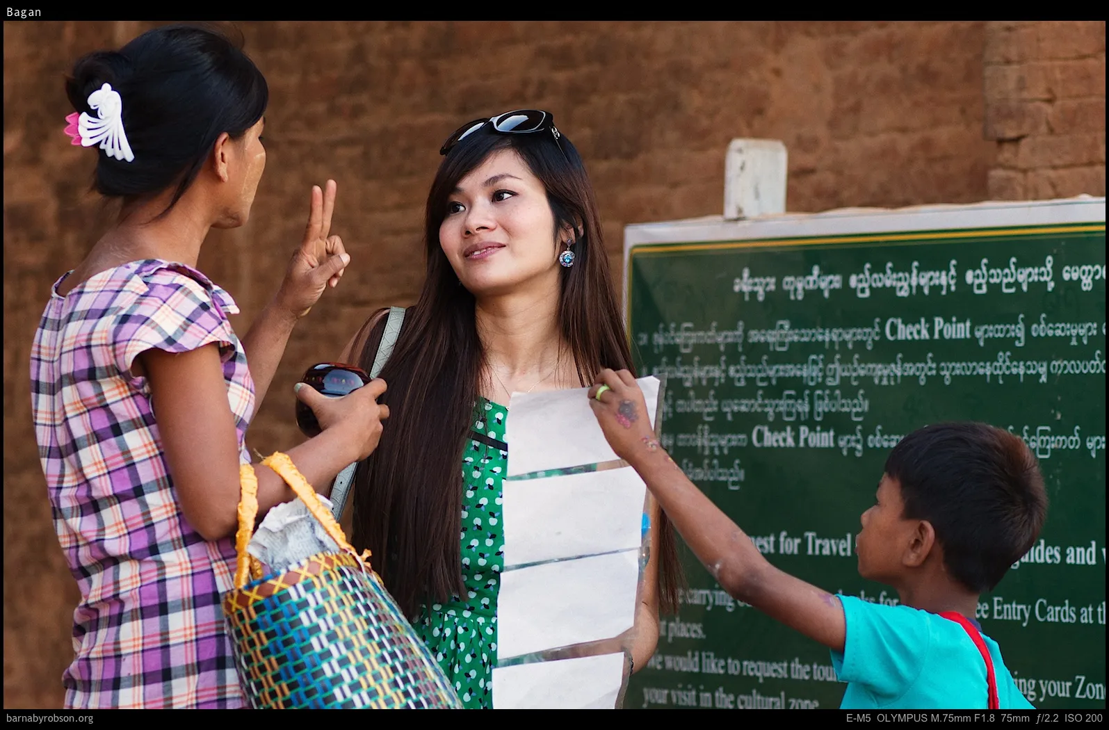 Woman gestures, another holds papers, child reaches for them near a green sign.