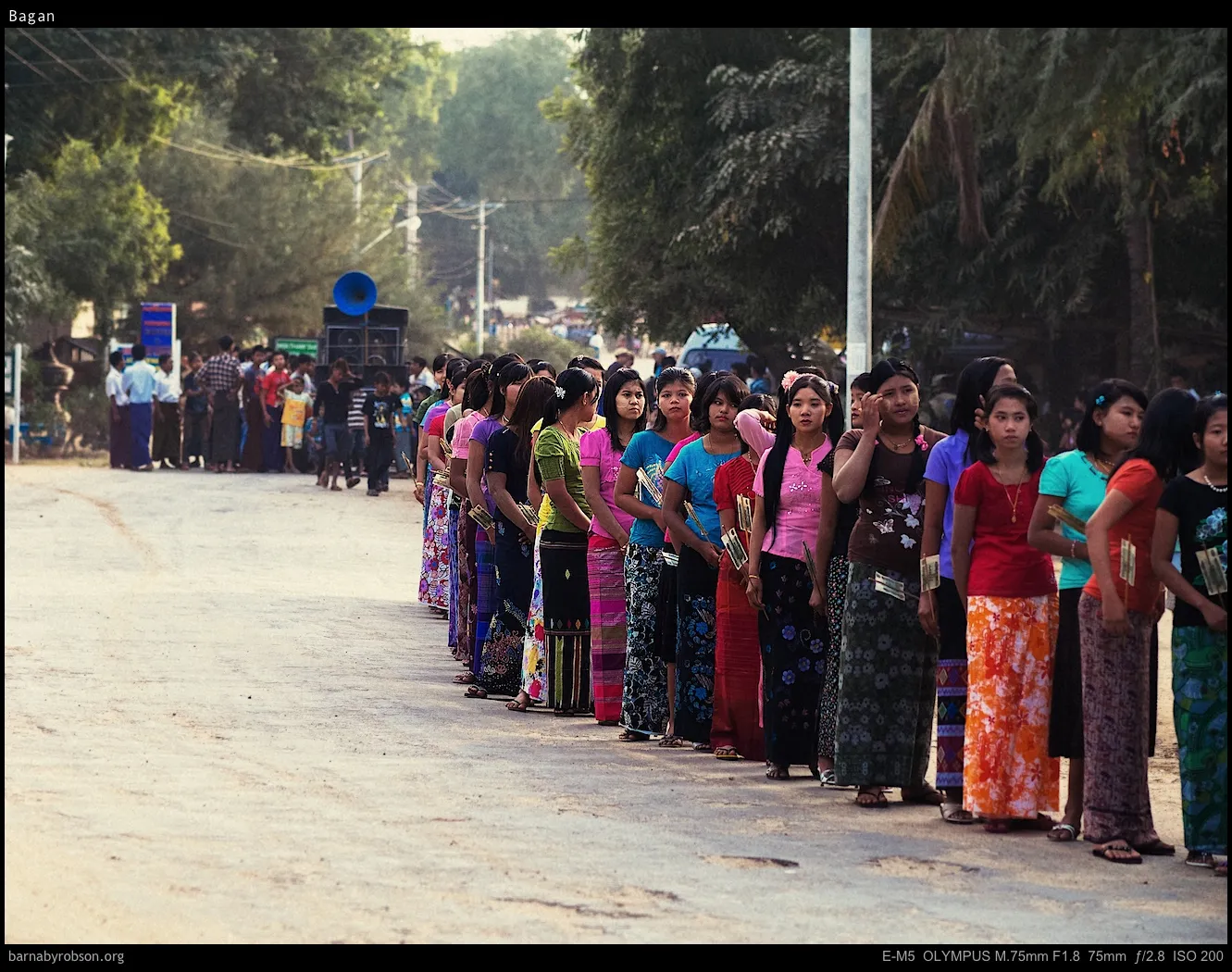 Queue of women in colourful traditional dress on a dusty road, blue speaker visible.