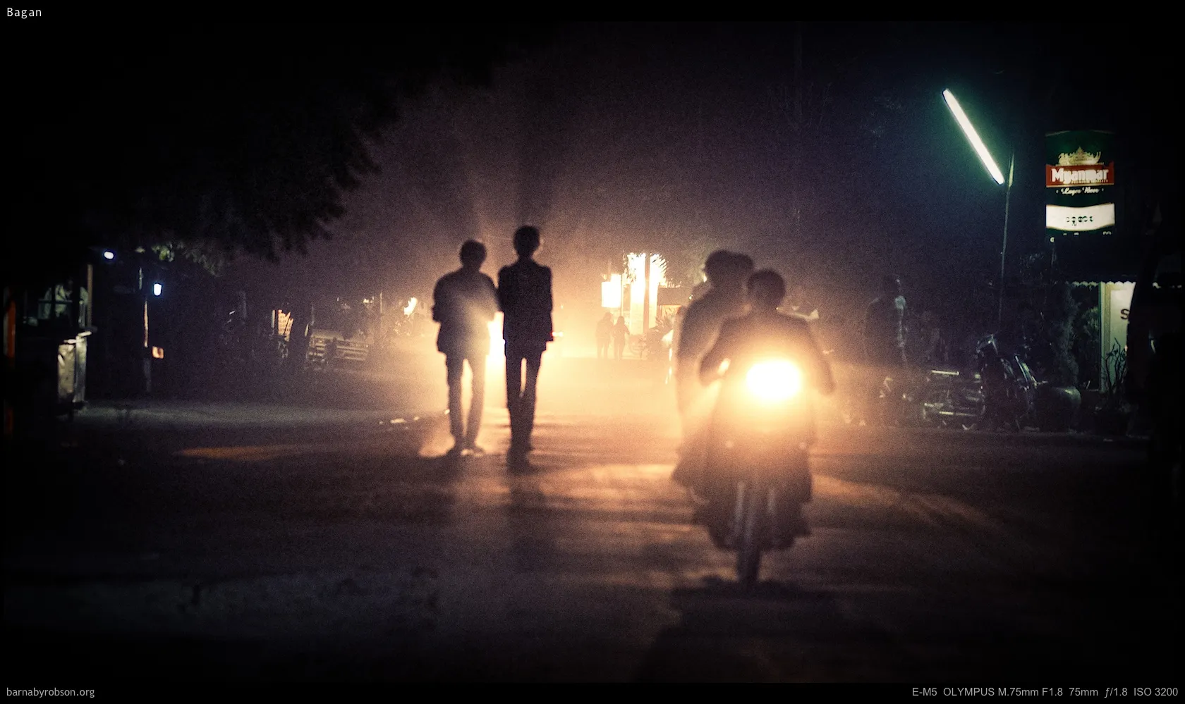 Night street with silhouetted figures, a motorbike, and bright hazy lights.