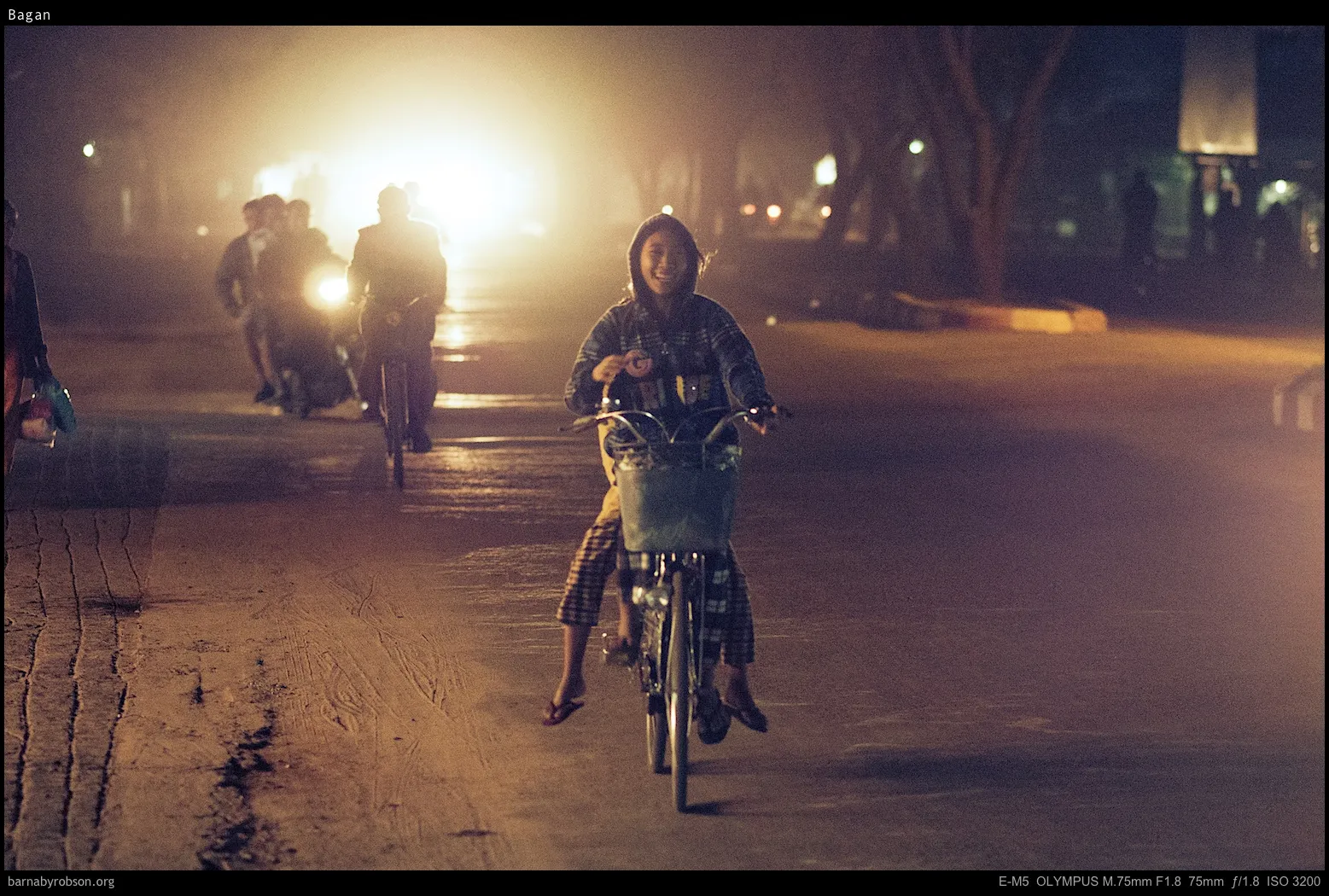 Smiling person cycling at night, other cyclists and bright lights behind.