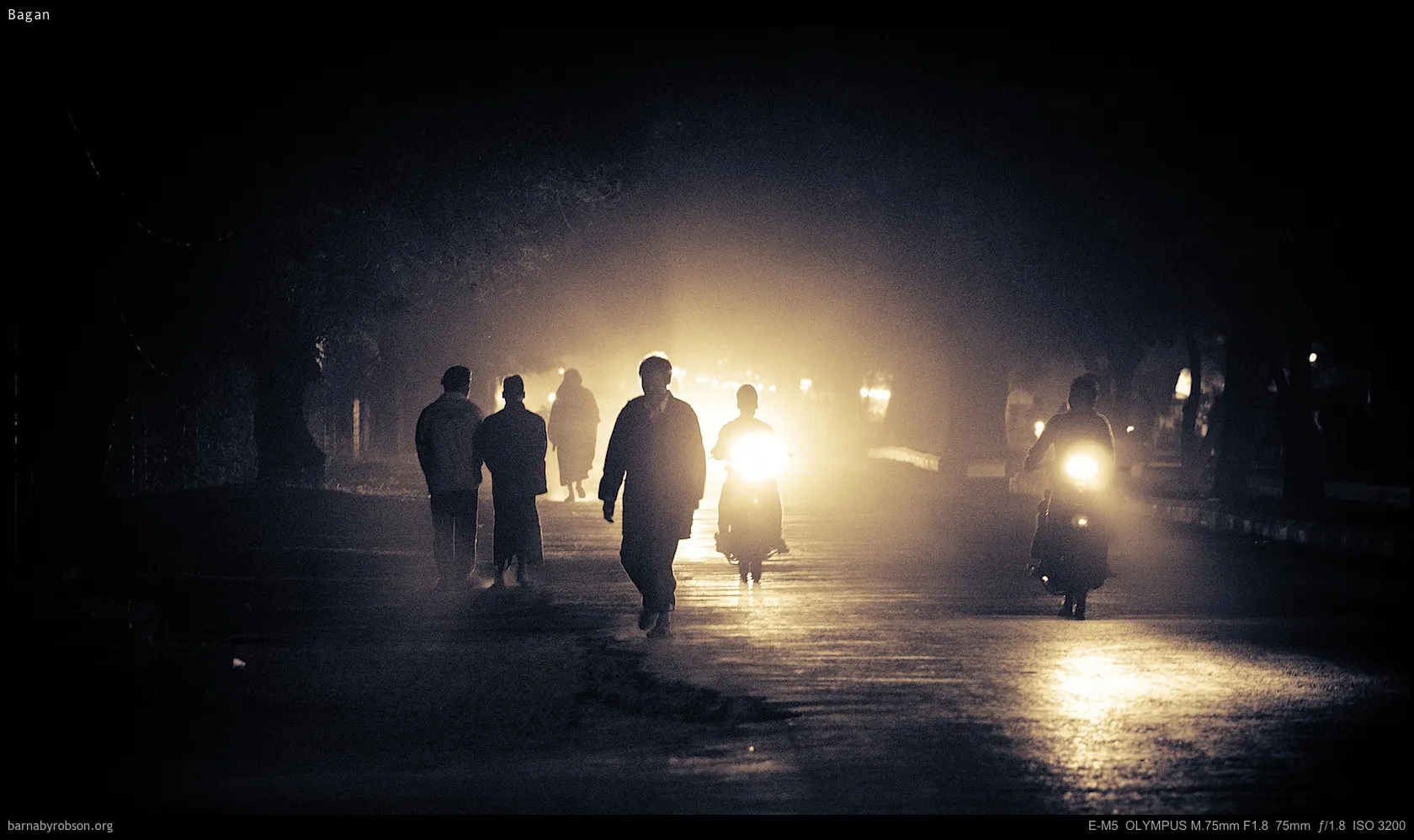 Pedestrians and motorbikes silhouetted by bright headlights on a dark, misty road.