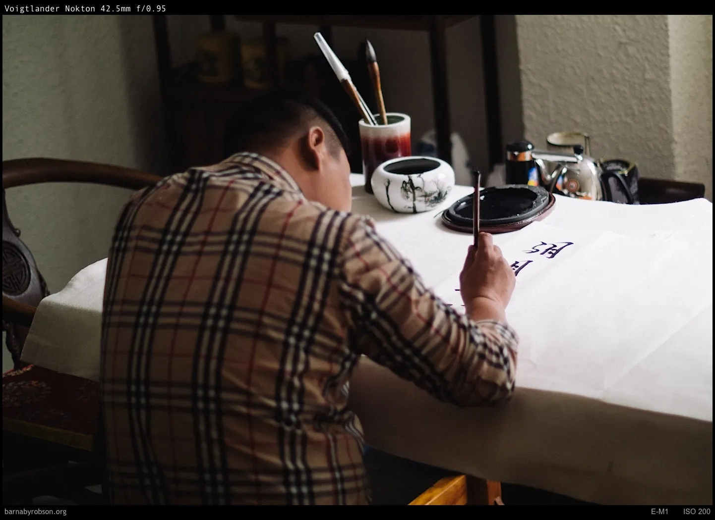 Person in checked shirt writing Chinese calligraphy with a brush.