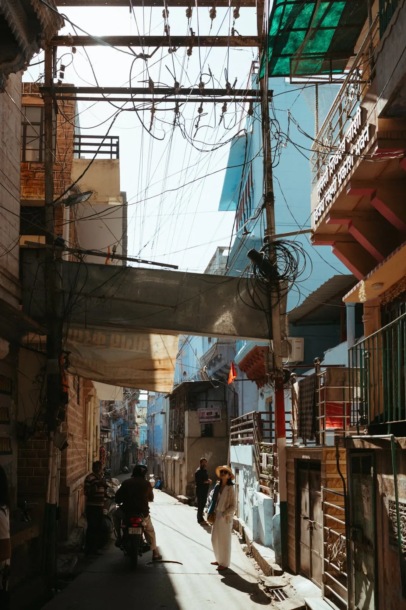 A narrow Jodhpur street with blue walls, tangled overhead wires, scooters and pedestrians in hard sunlight.