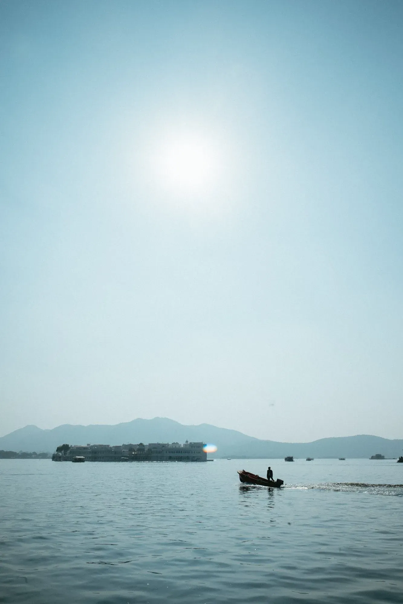 A small motorboat crossing Lake Pichola below a bright hazy sun and distant hills.