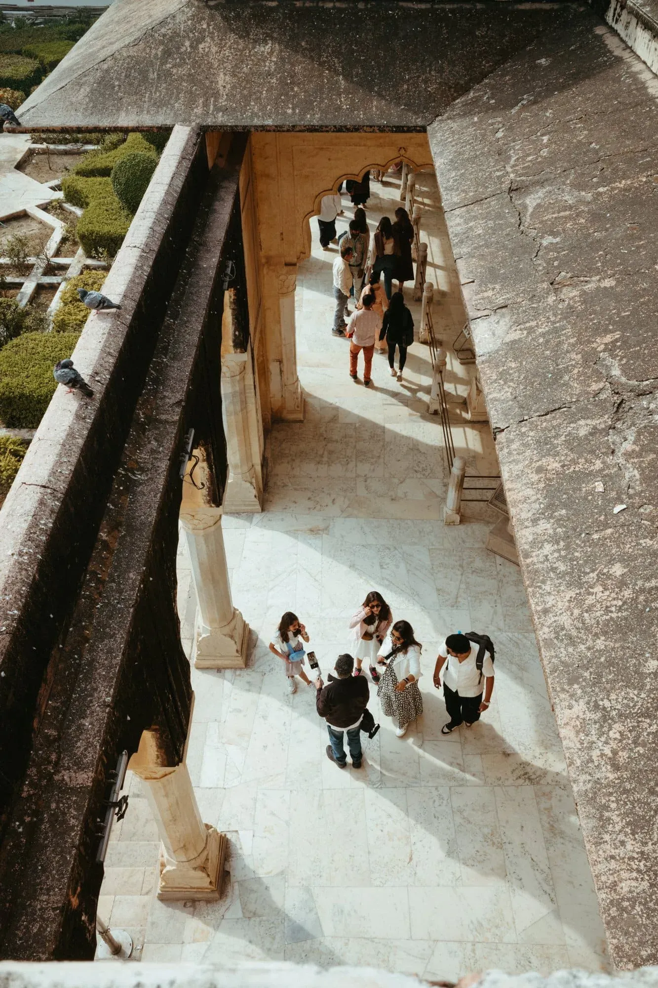 A high vertical view of visitors walking through a sunlit marble colonnade at Amber Fort.