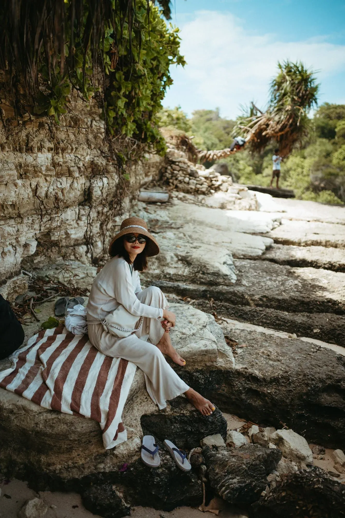 A woman in pale clothes and a wide-brimmed hat seated on layered coastal rocks.