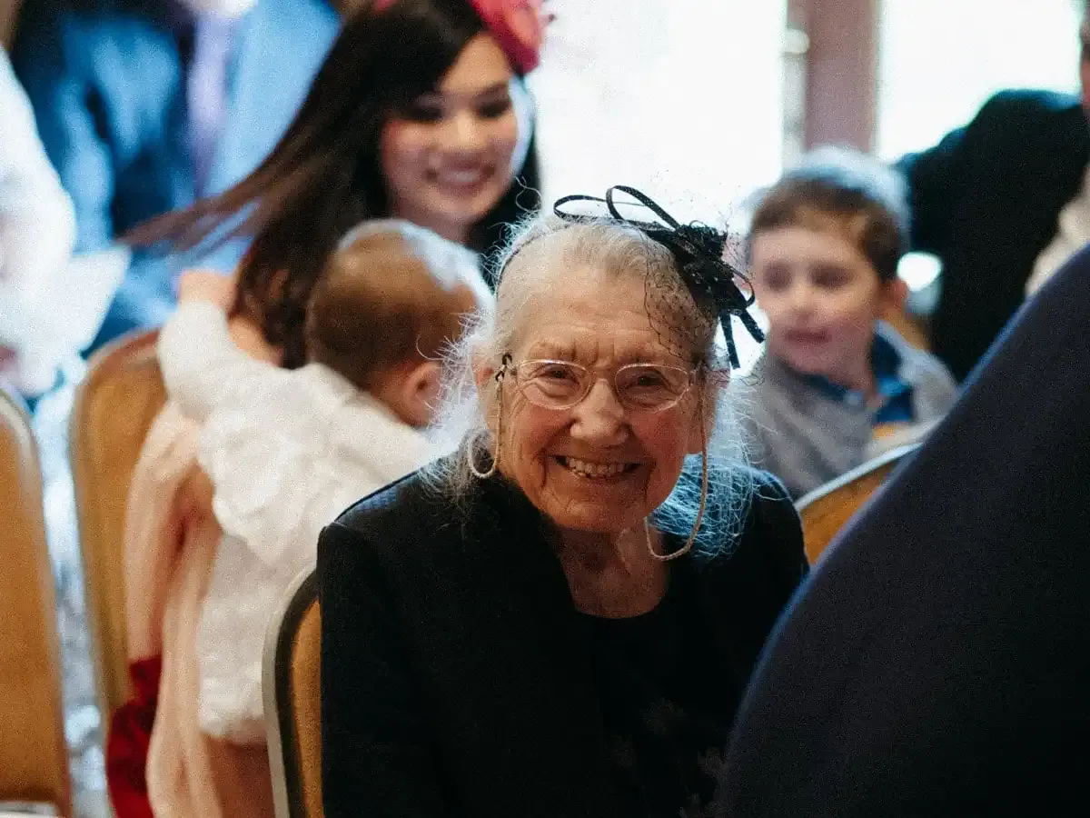 Smiling elderly woman in glasses and fascinator, with family in background.