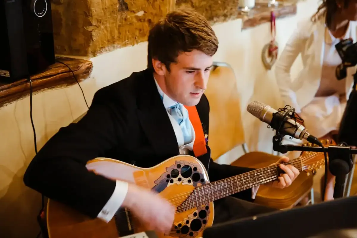 Man in black suit and blue cravat playing acoustic guitar into microphone.