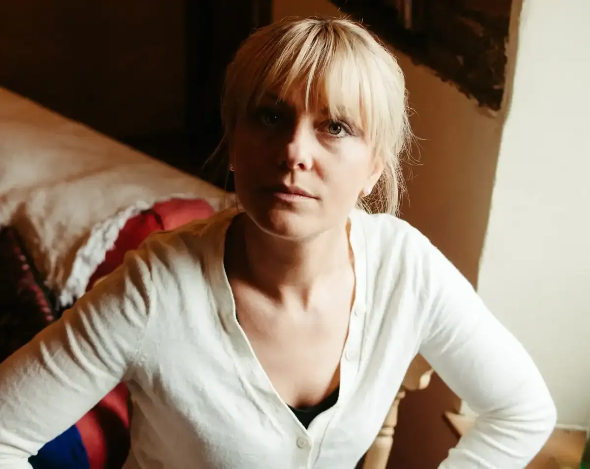 Woman with blonde fringe and white cardigan looking intently at camera.
