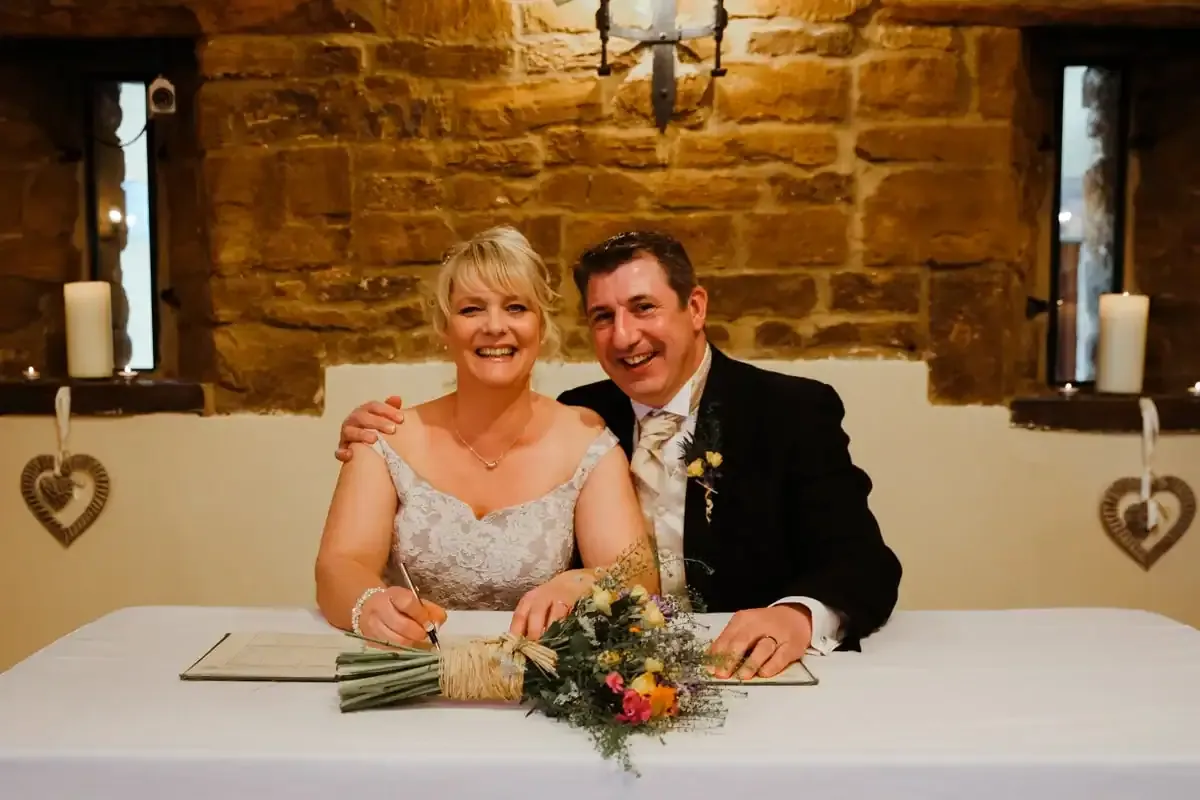 Bride signing wedding register, groom smiling beside her.