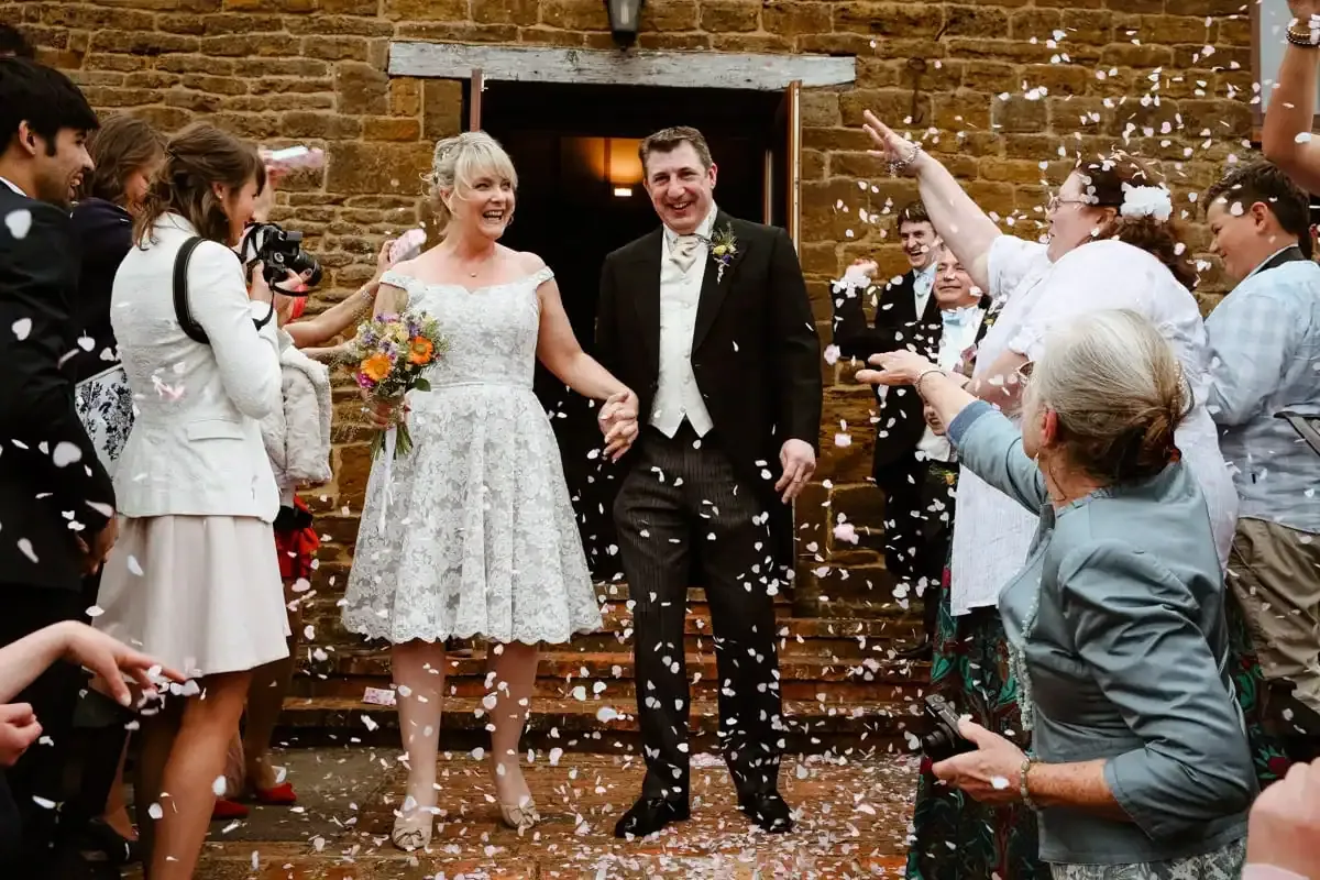 Bride and groom smiling, showered with confetti by guests outside a stone building.