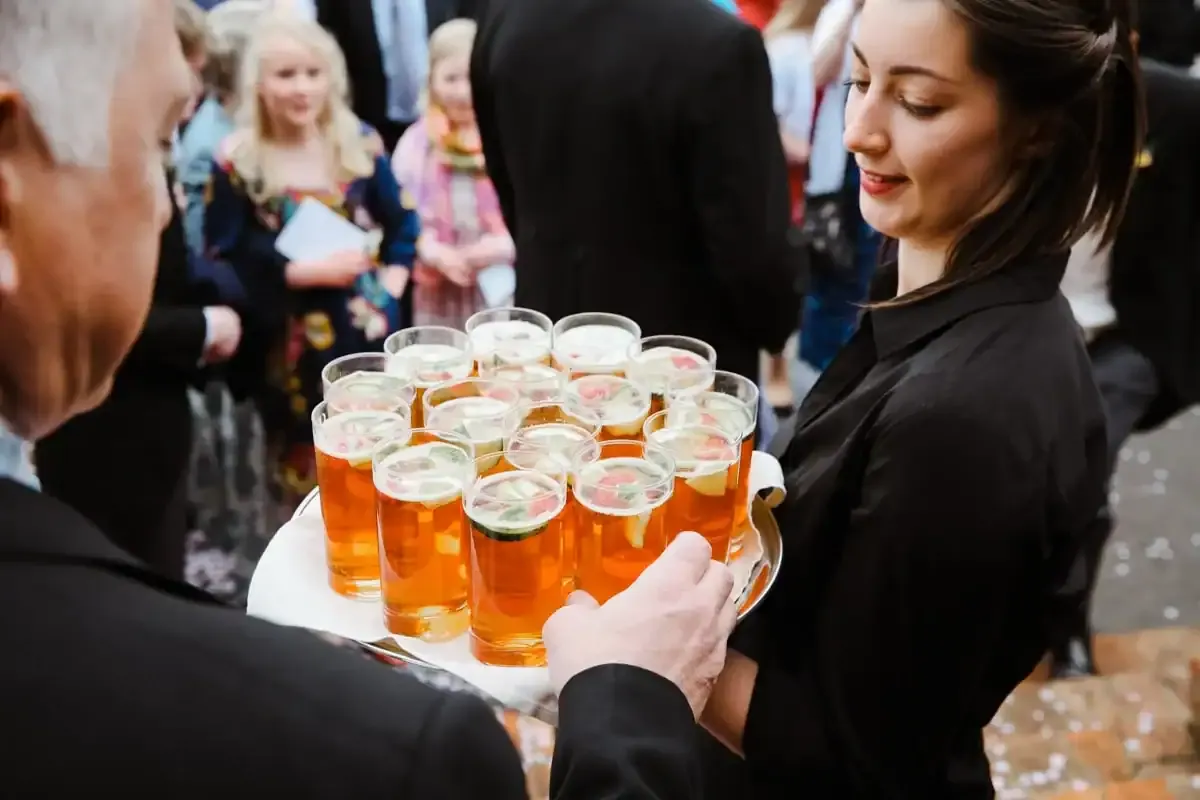 Woman serving multiple amber drinks with fruit and cucumber on a tray.