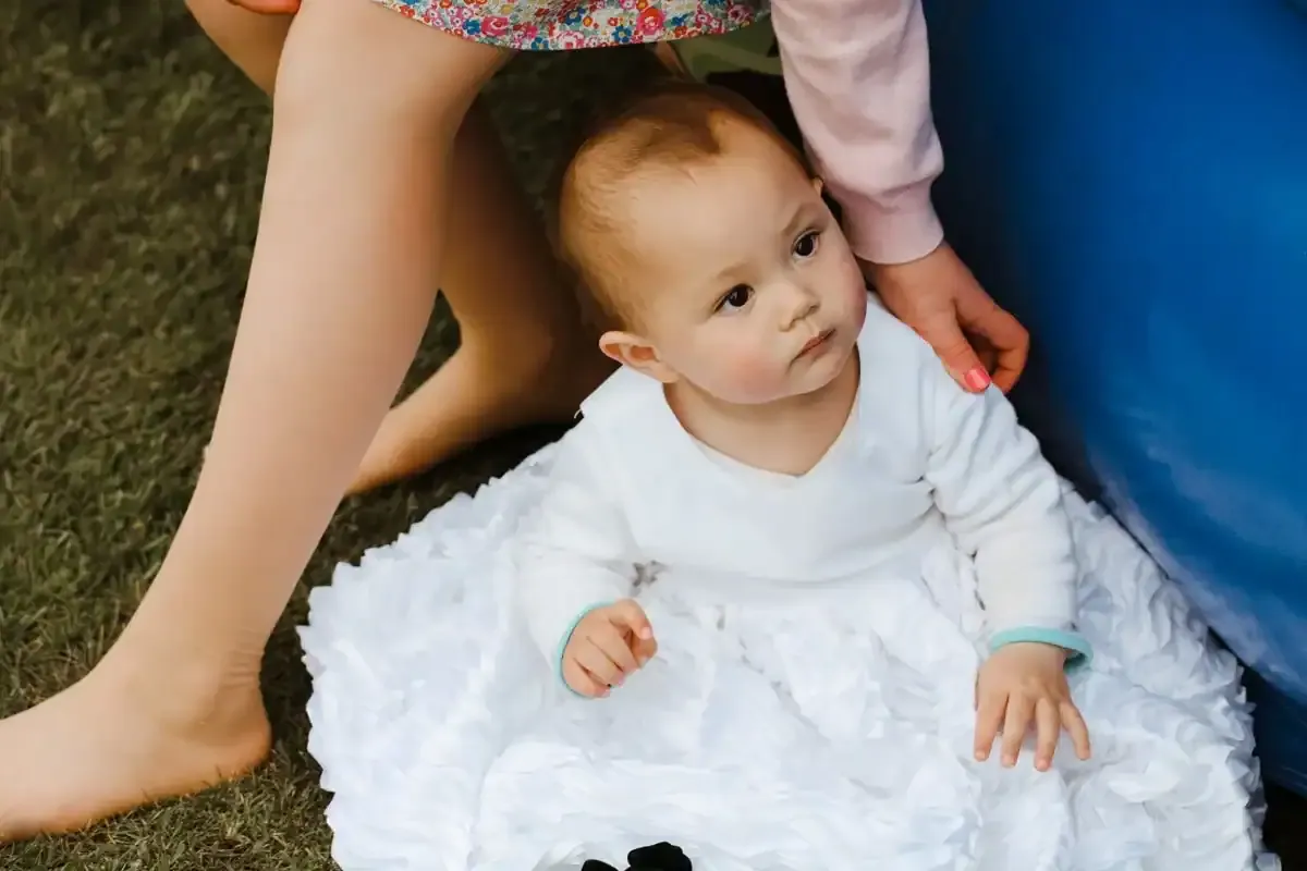 Baby in white ruffled dress on grass, adult's legs, hand, and blue object.