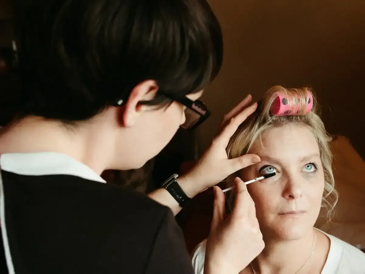 Makeup artist applies mascara to blonde woman with pink hair rollers.